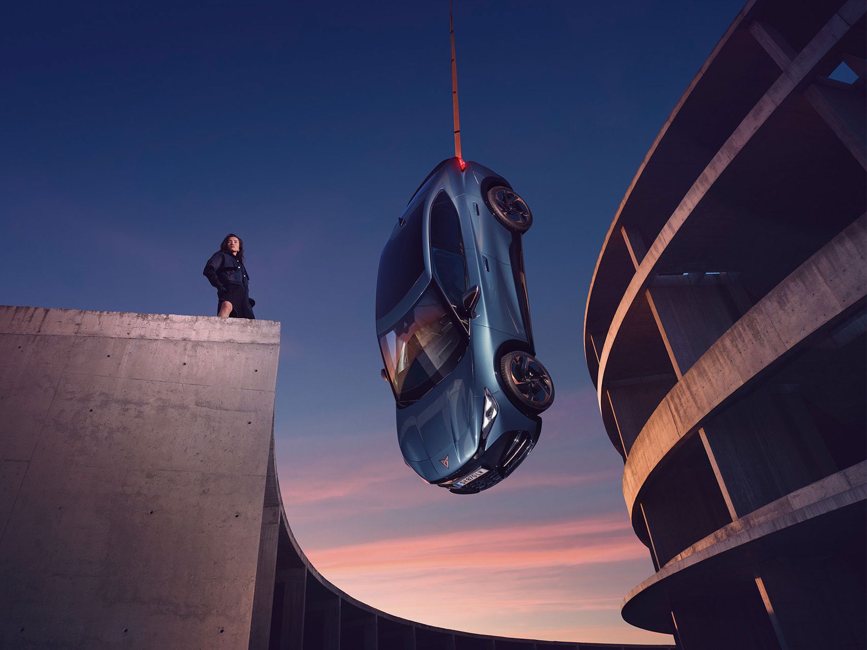 Blauer CUPRA Tavascan erscheint aufgehängt, eine Frau steht daneben auf einem Gebäude.