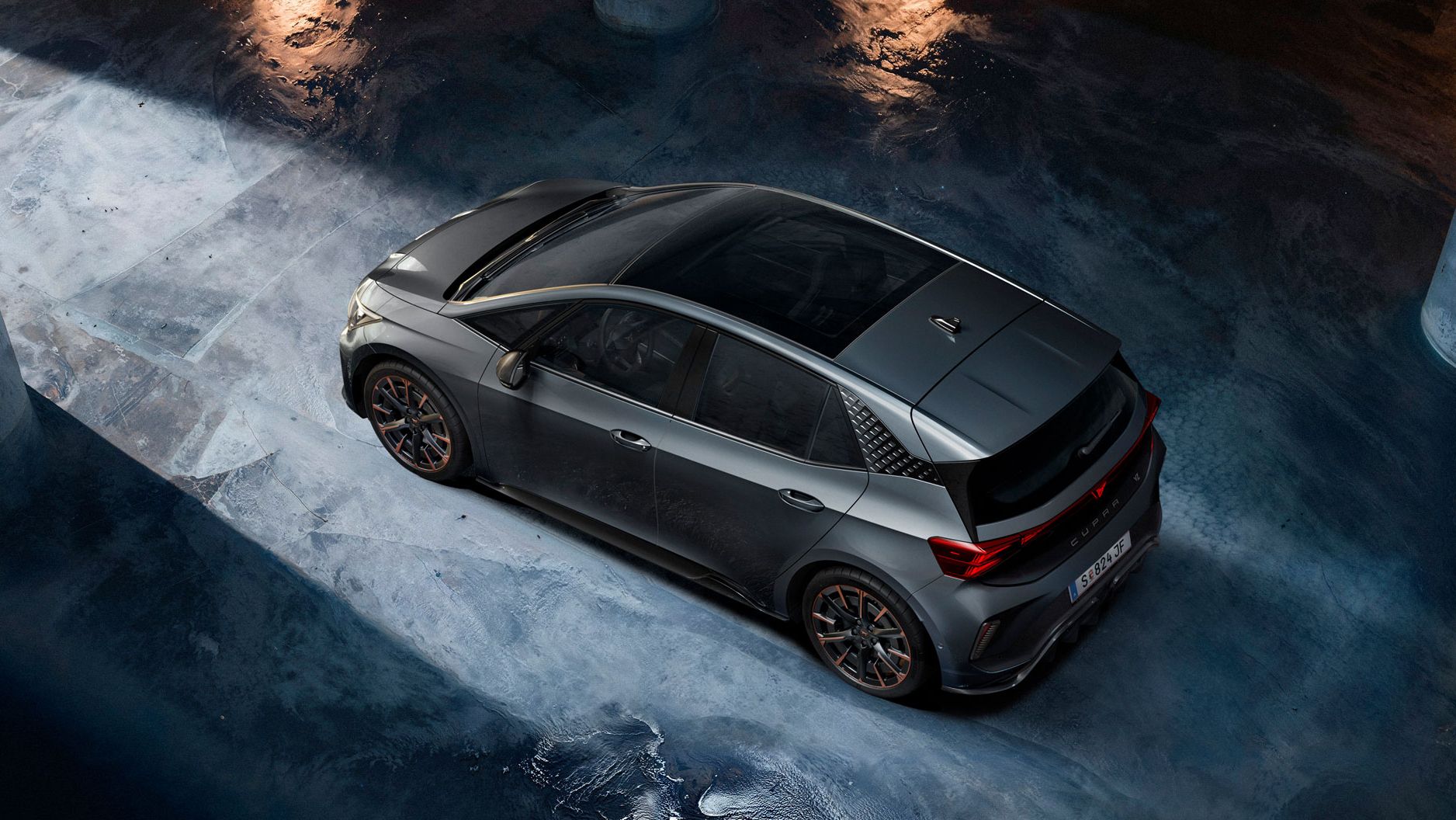 Ein grauer CUPRA Born steht in einer dunklen Tiefgarage. Das Bild ist aus der Vogelperspektive aufgenommen und zeigt Lichterscheinungen auf dem nassen Betonboden. Die Szenerie ist durch indirektes Licht beleuchtet, das eine dramatische Atmosphäre schafft.
