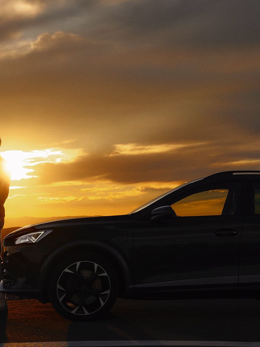 Ein Mann steht während des Sonnenuntergangs vor seinem CUPRA Formentor und blickt auf sein Smartphone.