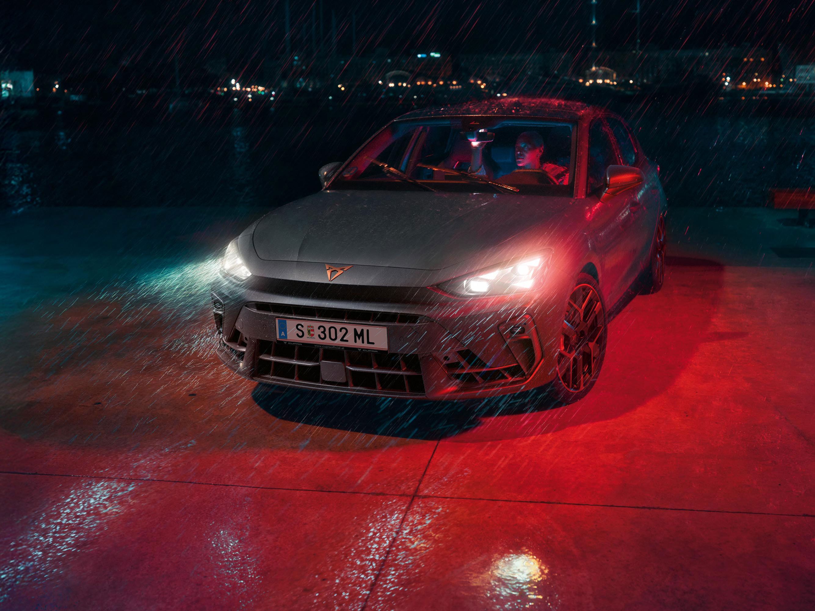 Ein Mann parkt seinen CUPRA Leon bei Nacht und Regenwetter an einem Gewässer. Im Hintergrund die Lichter einer Stadt.