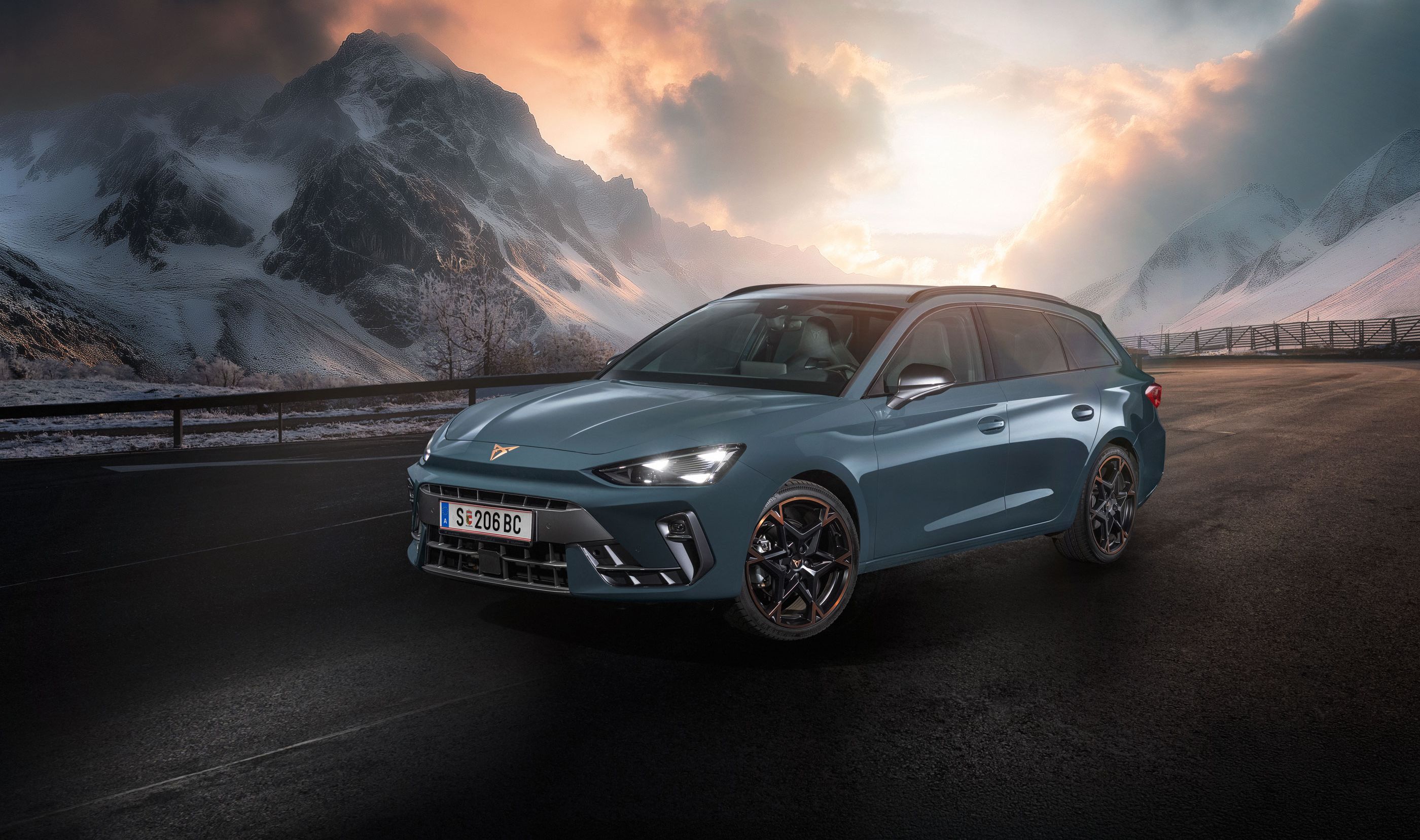 Ein CUPRA Leon Kombi parkt vor einer malerischen Berglandschaft bei Sonnenuntergang. Im Hintergrund sind verschneite Gipfel und dramatische Wolken zu sehen. Der Asphalt ist dunkel und feucht.