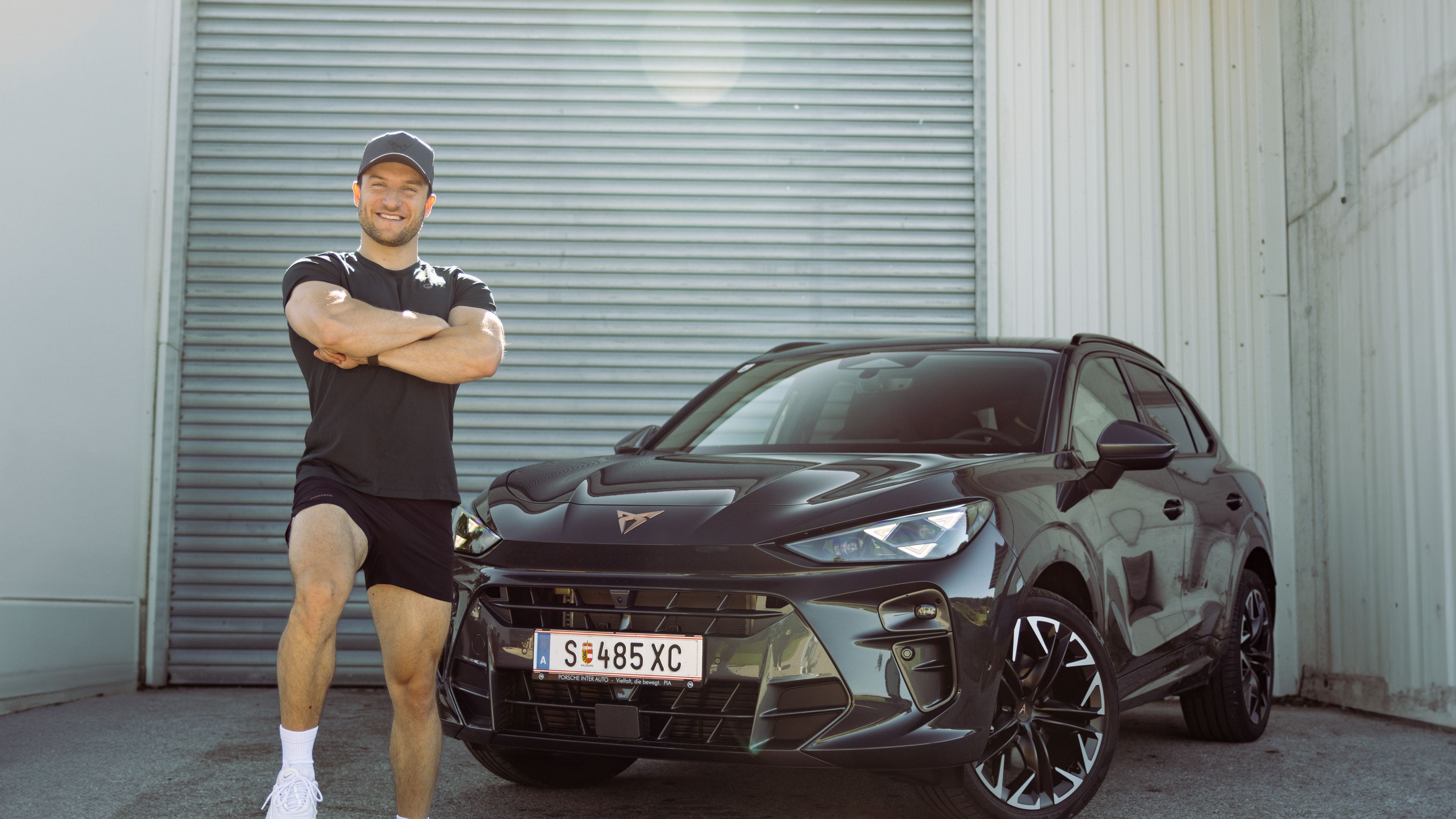 Sascha Huber vor Auto mit Fuß auf Hantel abgestellt und Arme verschränkt
