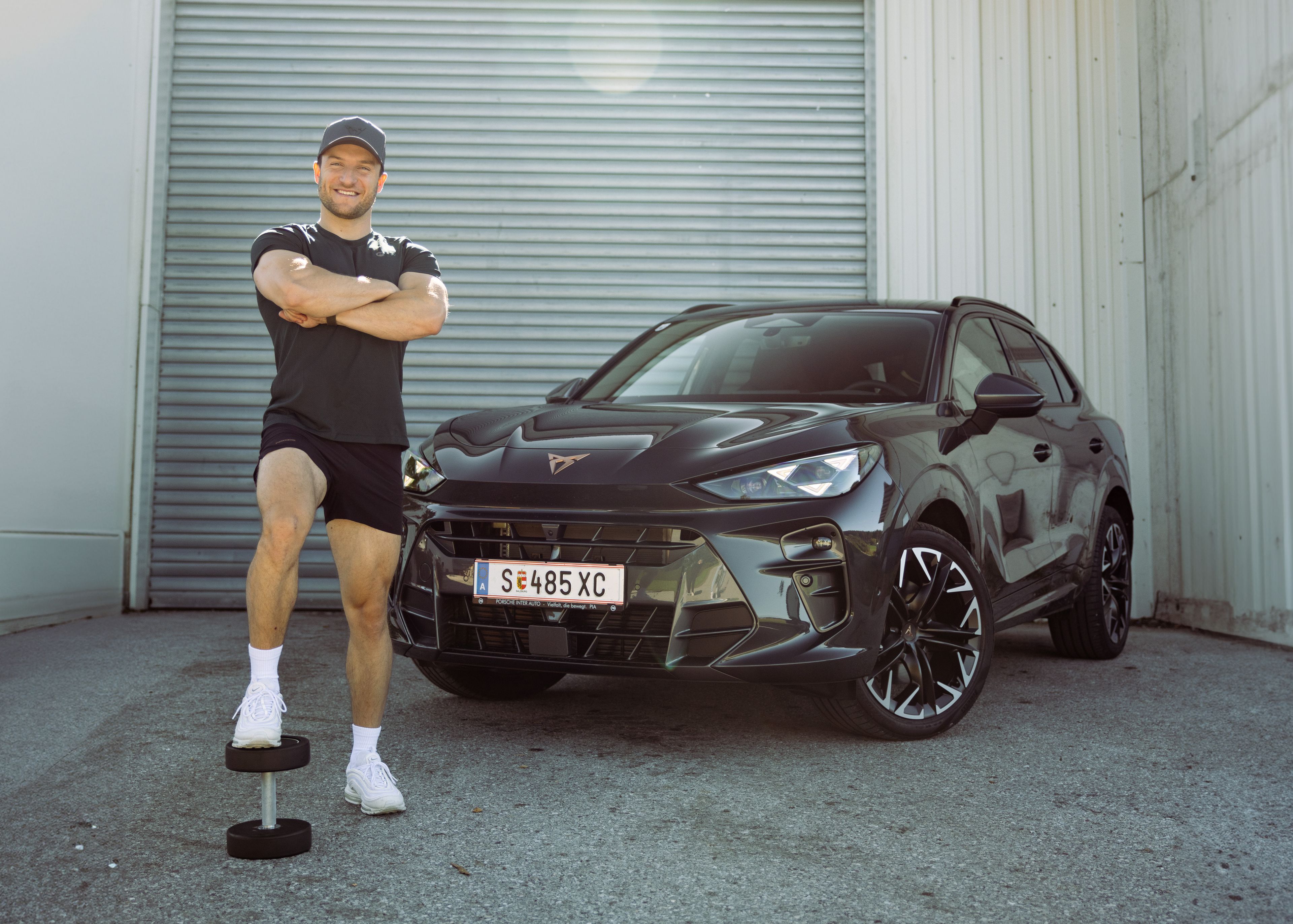 Sascha Huber vor Auto mit Fuß auf Hantel abgestellt und Arme verschränkt