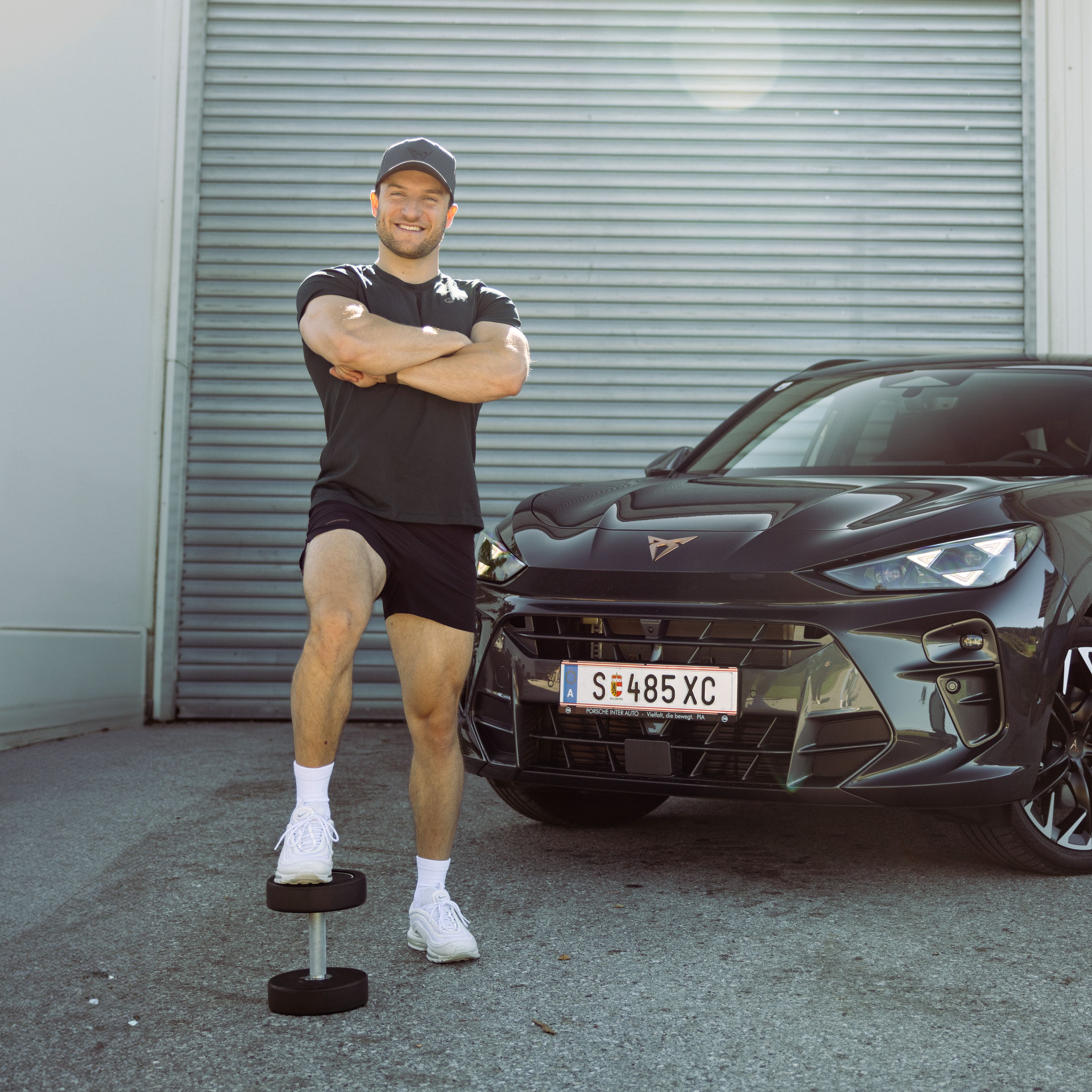 Sascha Huber vor Auto mit Fuß auf Hantel abgestellt und Arme verschränkt