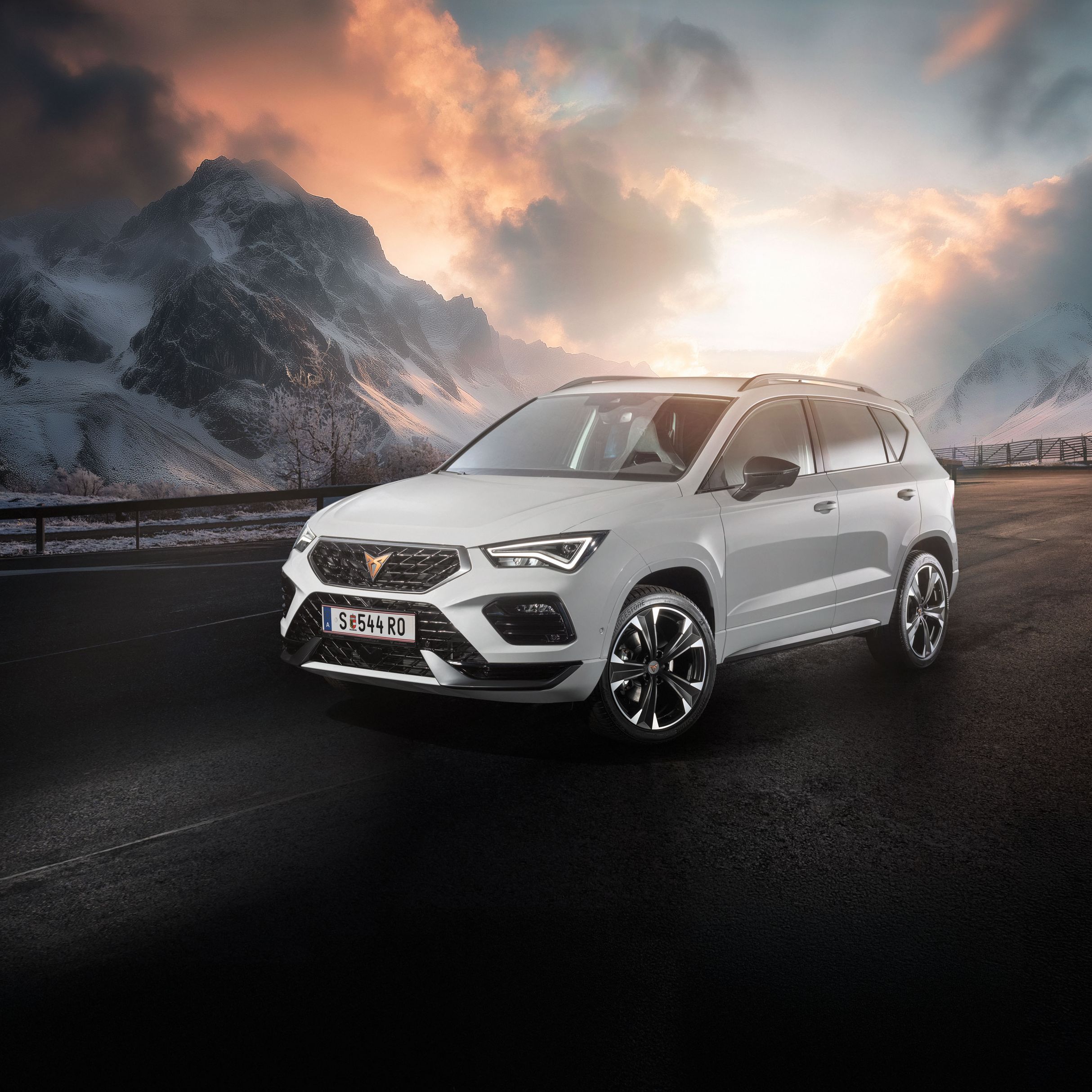 Ein weißer CUPRA Ateca fährt auf einer Bergstraße bei Sonnenaufgang. Die Straße ist nass und von Schnee gesäumt. Im Hintergrund sind schneebedeckte Berge und bewölkter Himmel zu sehen.