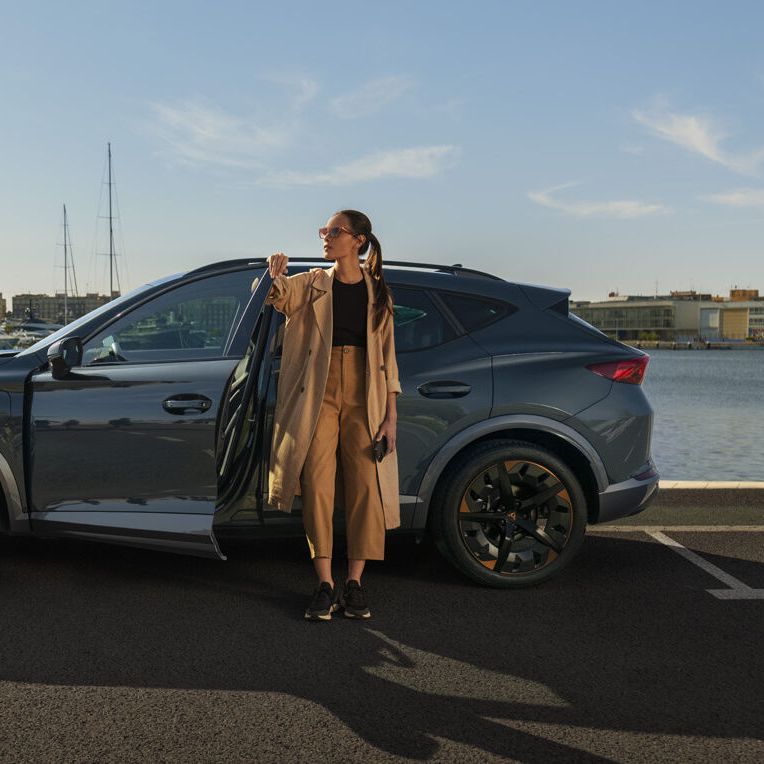 Eine Frau steht an der geöffneten Vordertüre eines grauen CUPRA Leon der am Kai eines Hafens parkt.