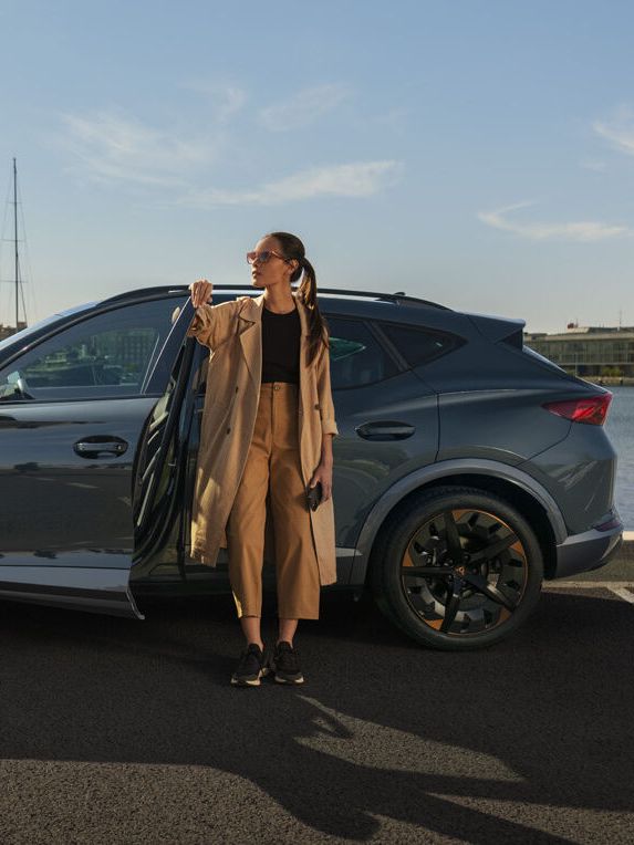 Eine Frau steht an der geöffneten Vordertüre eines grauen CUPRA Leon der am Kai eines Hafens parkt.