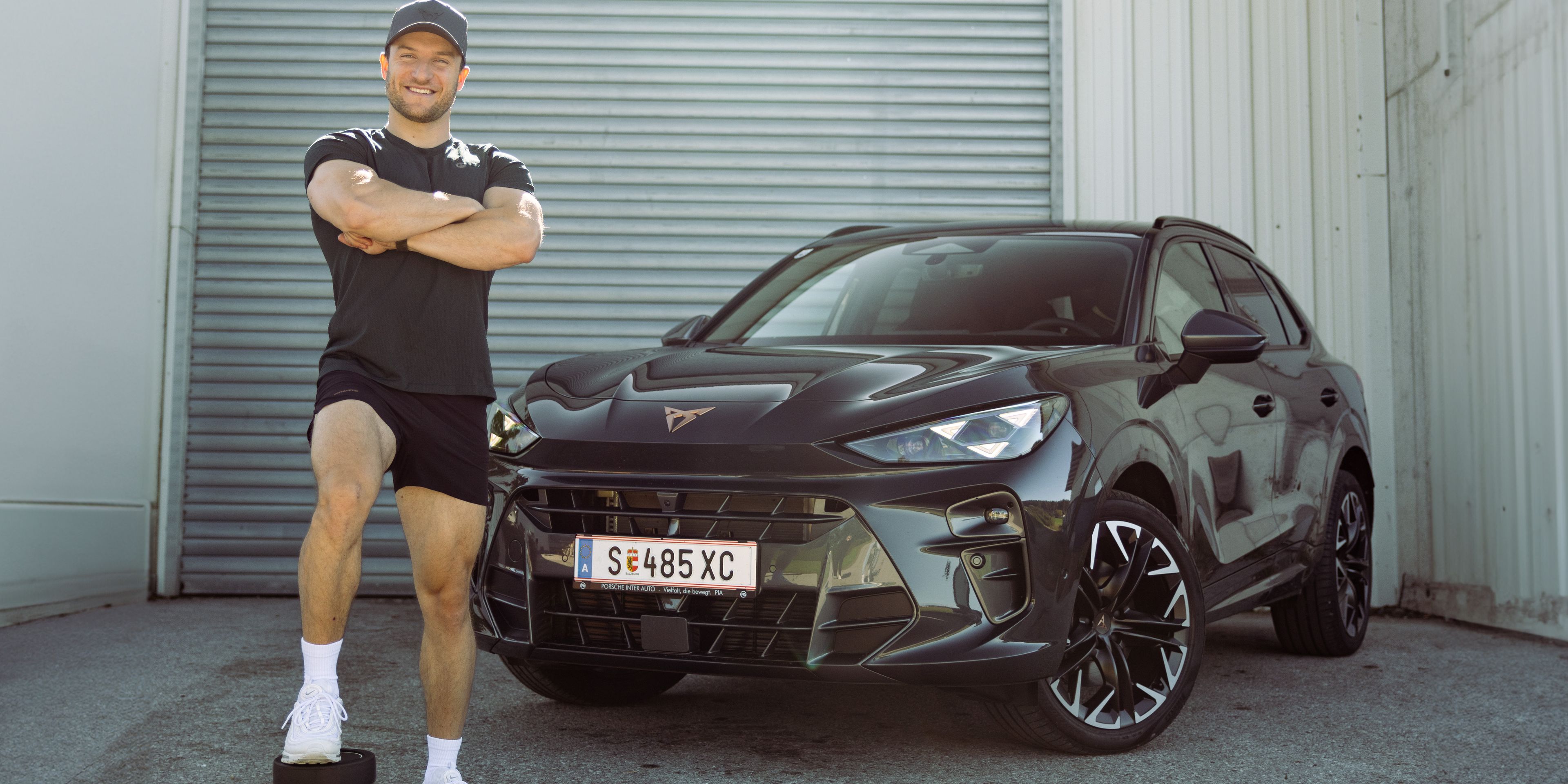 Sascha Huber vor Auto mit Fuß auf Hantel abgestellt und Arme verschränkt