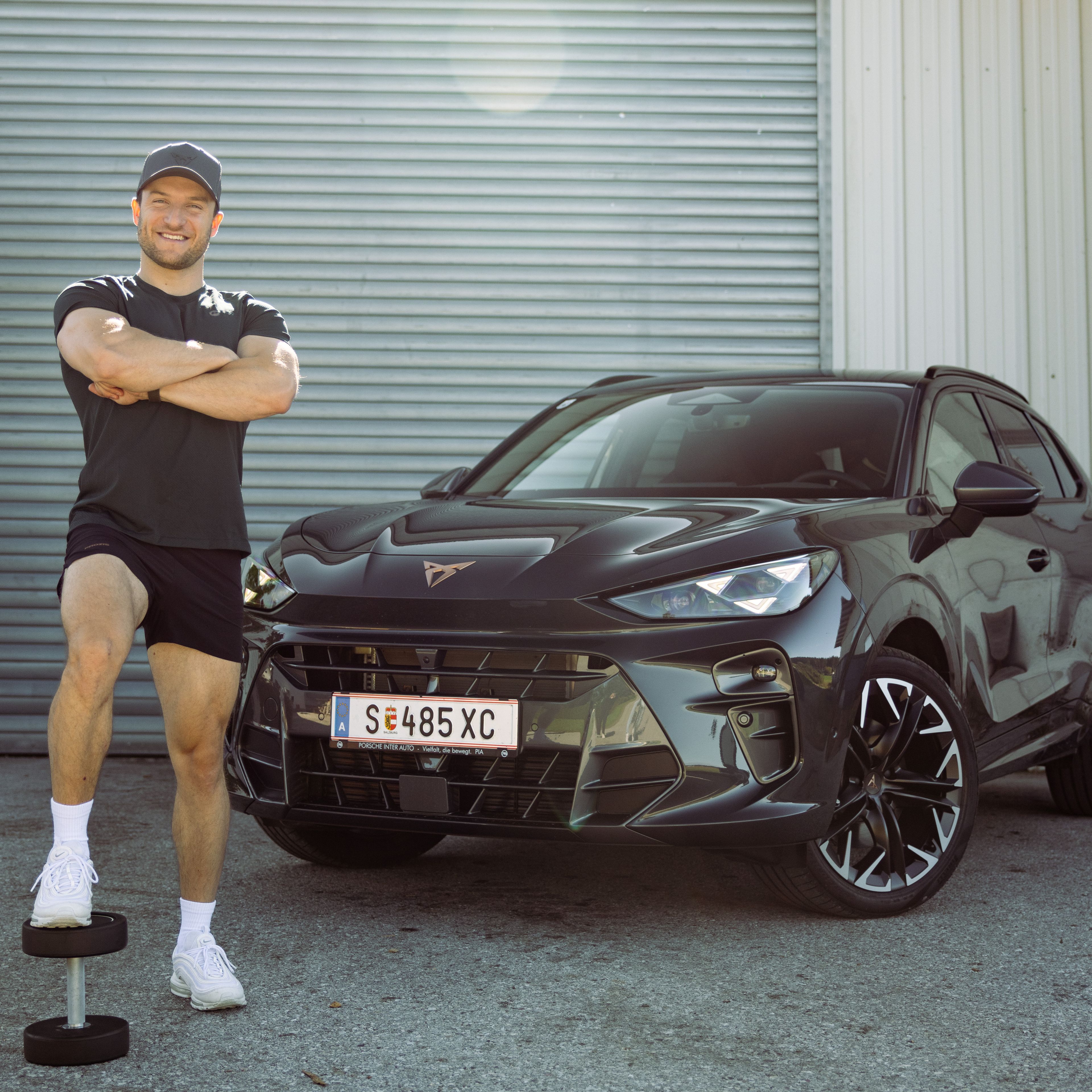 Sascha Huber vor Auto mit Fuß auf Hantel abgestellt und Arme verschränkt