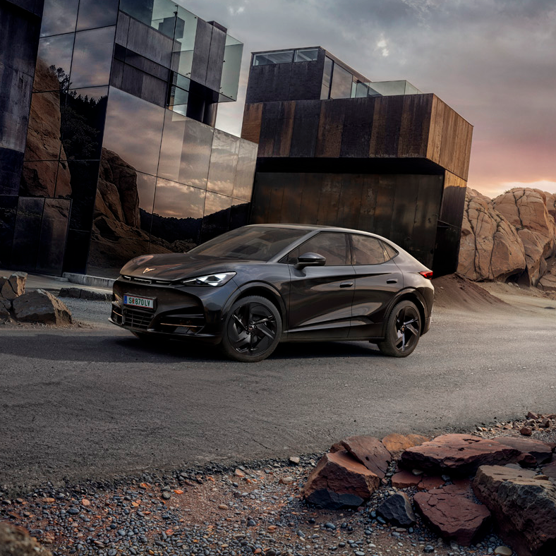 CUPRA Tavascan in schwarz mit schwarzen Felgen vor Glasgebäude und Felsen geparkt