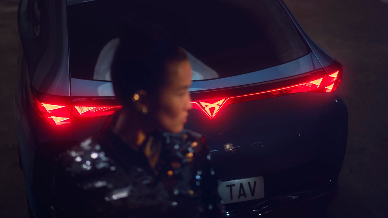 Eine Frau steht hinter einem CUPRA Tavascan mit eingeschalteten Rückleuchten.. 