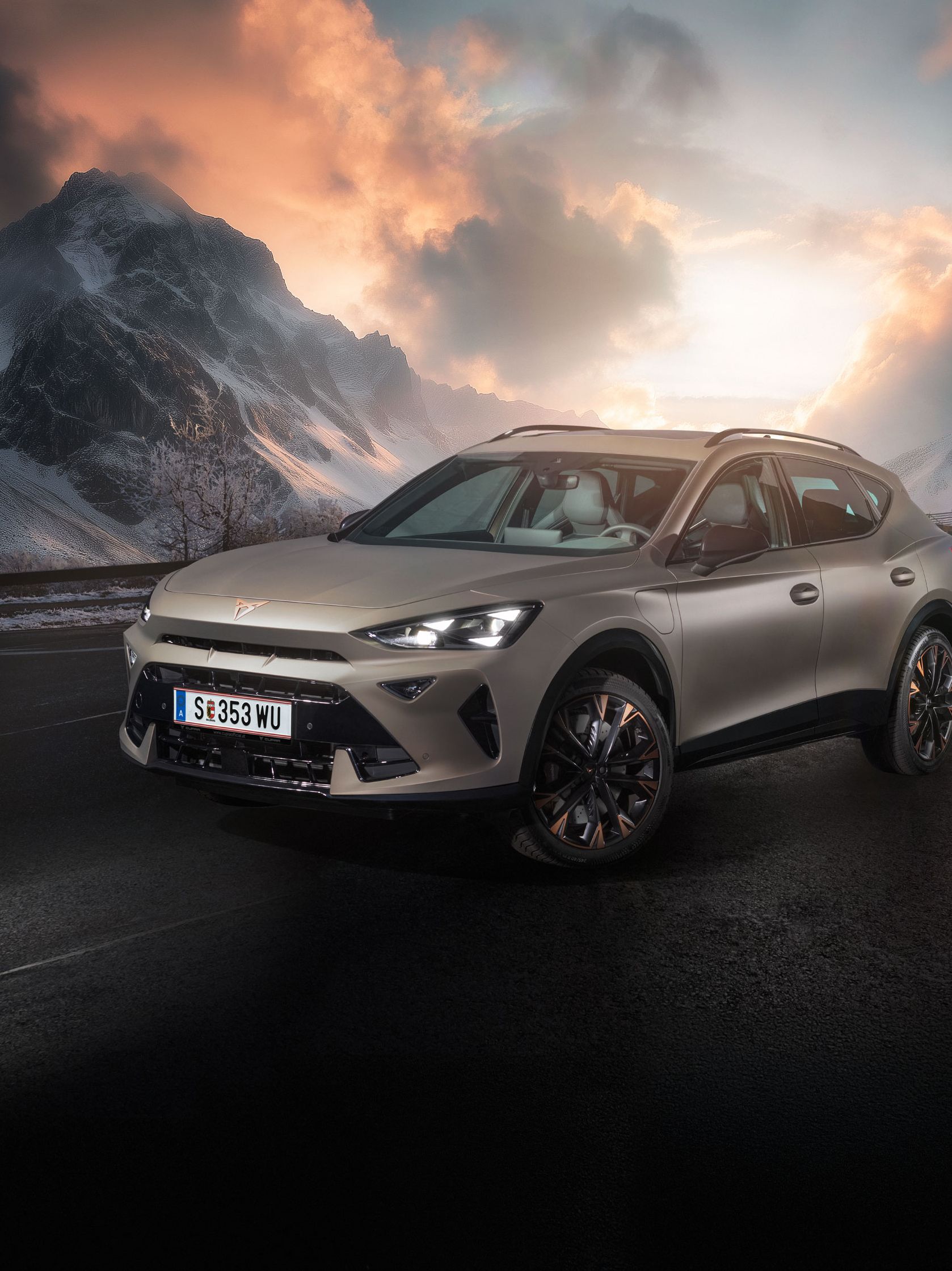 Ein CUPRA Formentor steht auf einer Straße in bergiger Landschaft mit schneebedeckten Gipfeln. Der Himmel ist dramatisch bewölkt. Im Hintergrund scheint die Sonne durch die Wolken.