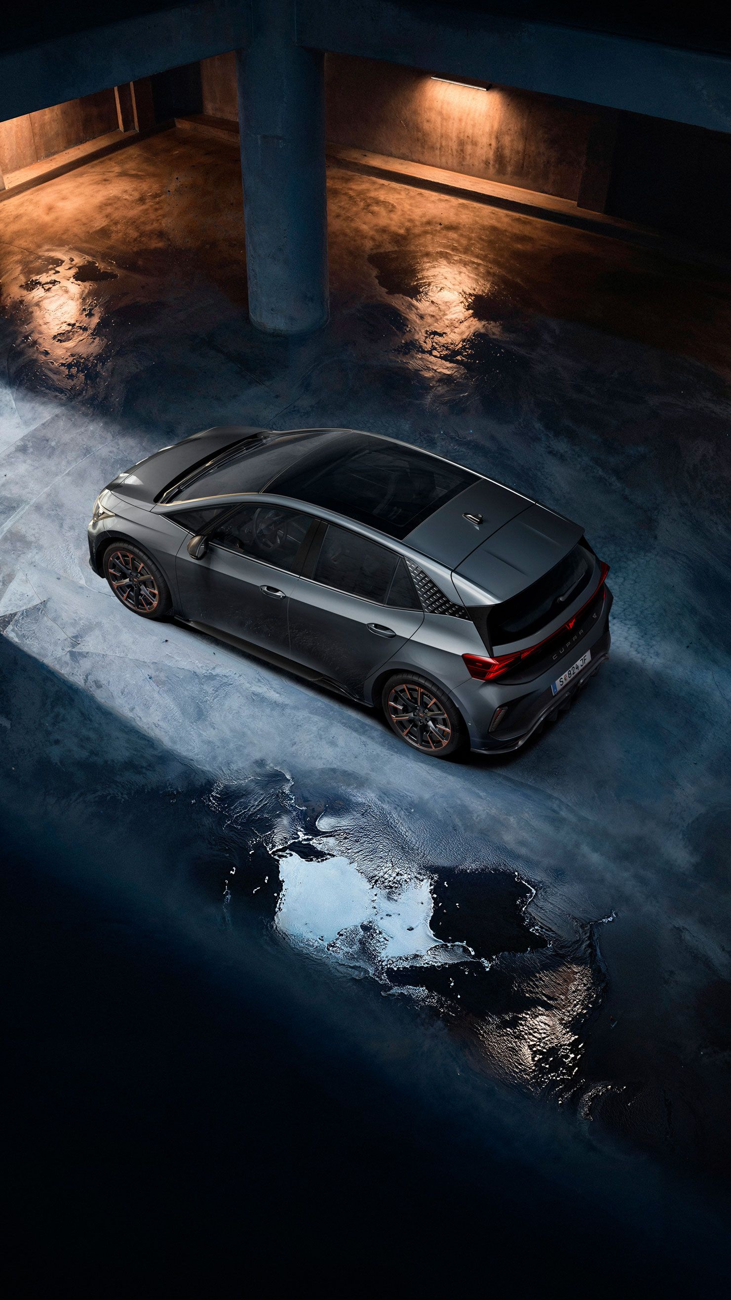 Ein grauer CUPRA Born steht in einer dunklen Tiefgarage. Das Bild ist aus der Vogelperspektive aufgenommen und zeigt Lichterscheinungen auf dem nassen Betonboden. Die Szenerie ist durch indirektes Licht beleuchtet, das eine dramatische Atmosphäre schafft.