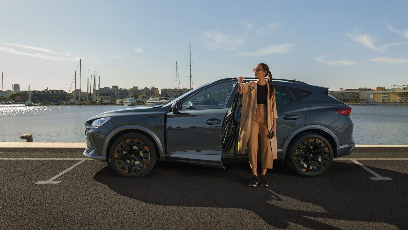 Eine Frau steht an der geöffneten Vordertüre eines grauen CUPRA Leon der am Kai eines Hafens parkt.