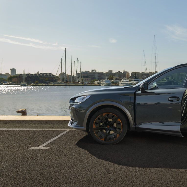 Eine Frau steht an der geöffneten Vordertüre eines grauen CUPRA Leon der am Kai eines Hafens parkt.
