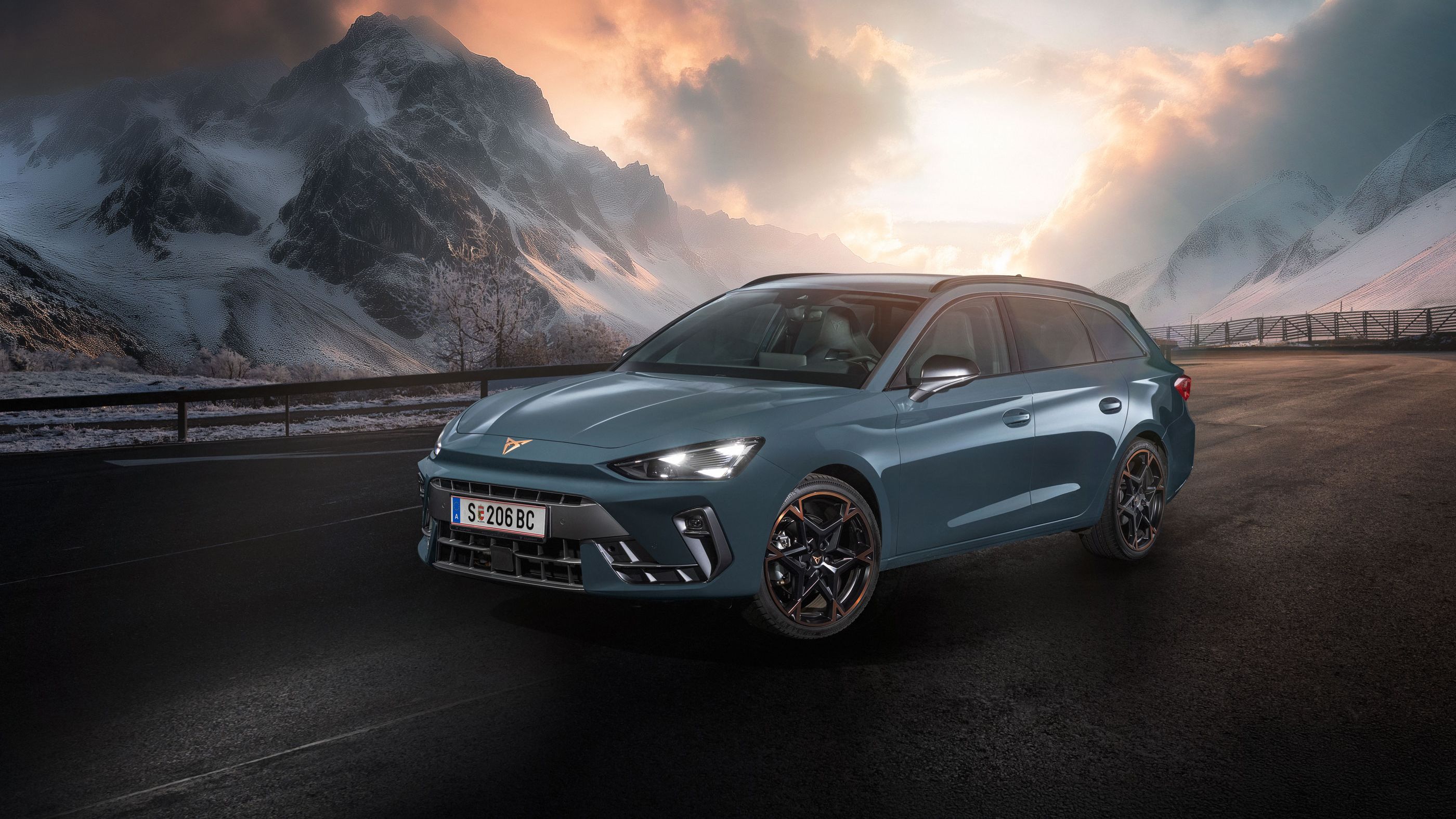 Ein CUPRA Leon Kombi parkt vor einer malerischen Berglandschaft bei Sonnenuntergang. Im Hintergrund sind verschneite Gipfel und dramatische Wolken zu sehen. Der Asphalt ist dunkel und feucht.