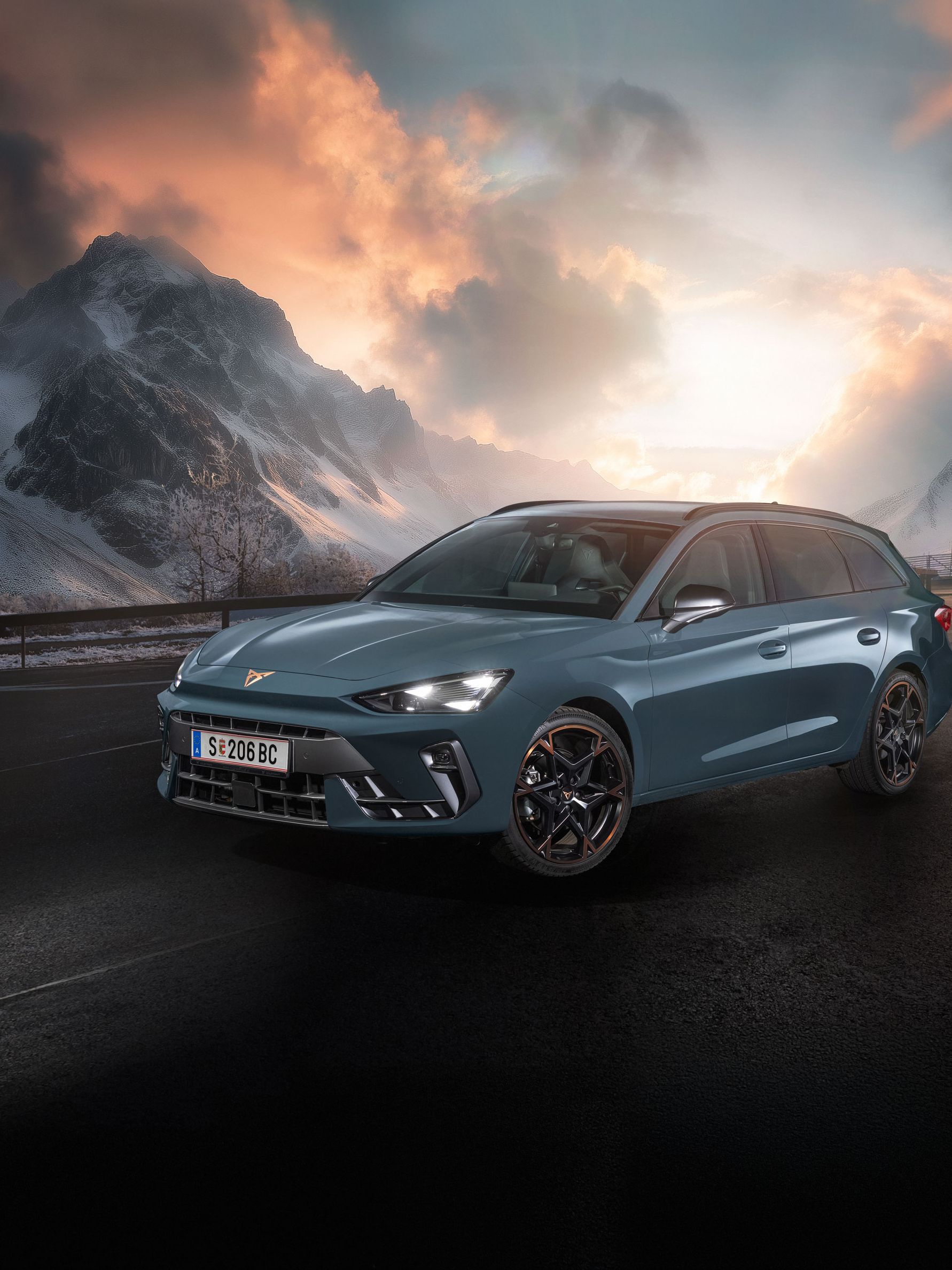 Ein CUPRA Leon Kombi parkt vor einer malerischen Berglandschaft bei Sonnenuntergang. Im Hintergrund sind verschneite Gipfel und dramatische Wolken zu sehen. Der Asphalt ist dunkel und feucht.