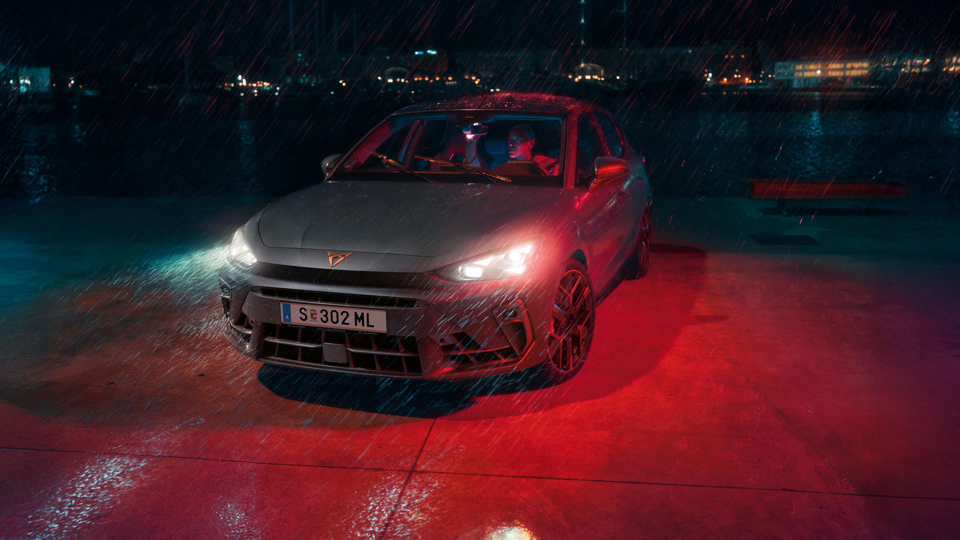 Ein Mann parkt seinen CUPRA Leon bei Nacht und Regenwetter an einem Gewässer. Im Hintergrund die Lichter einer Stadt.