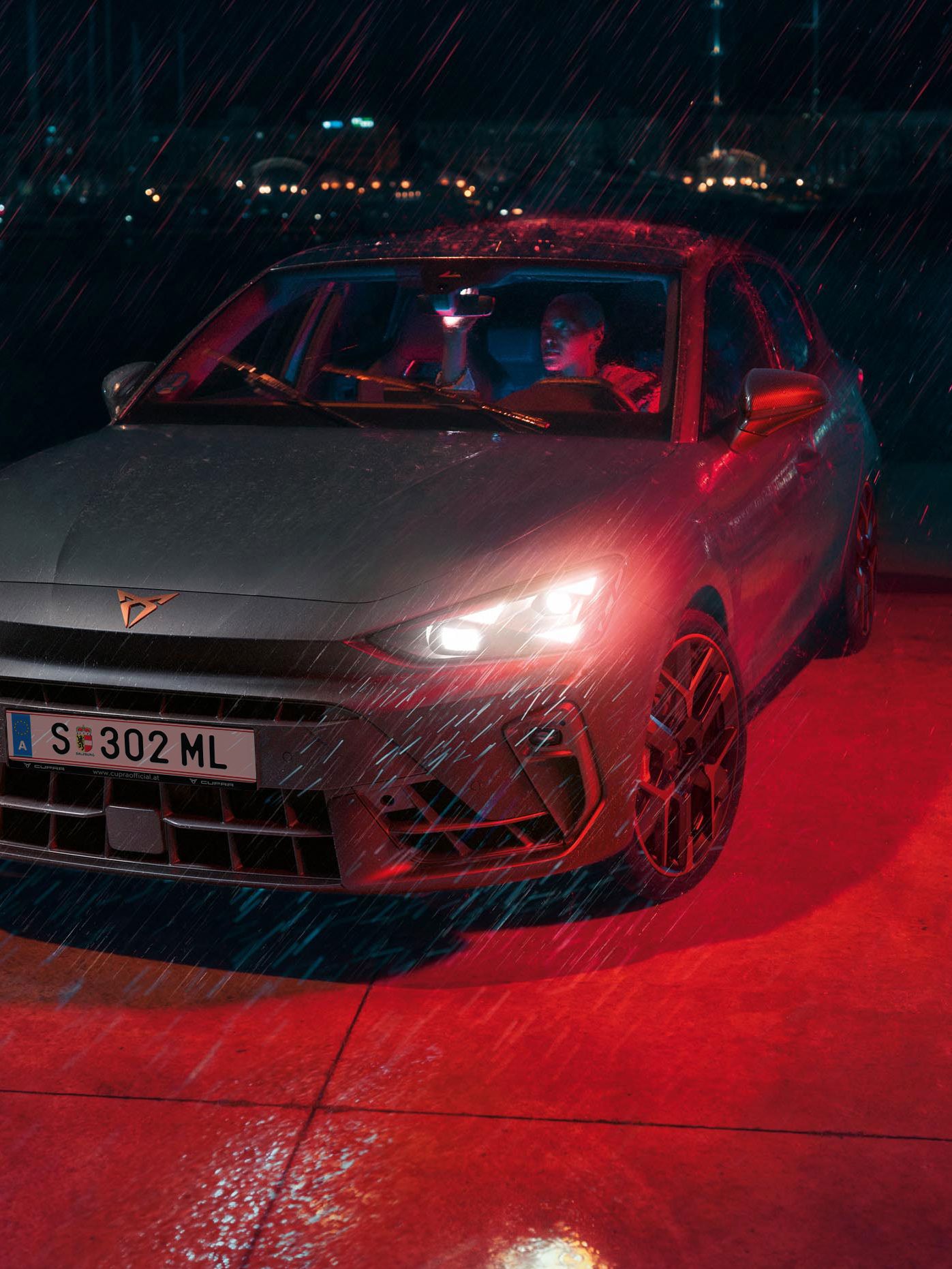 Ein Mann parkt seinen CUPRA Leon bei Nacht und Regenwetter an einem Gewässer. Im Hintergrund die Lichter einer Stadt.