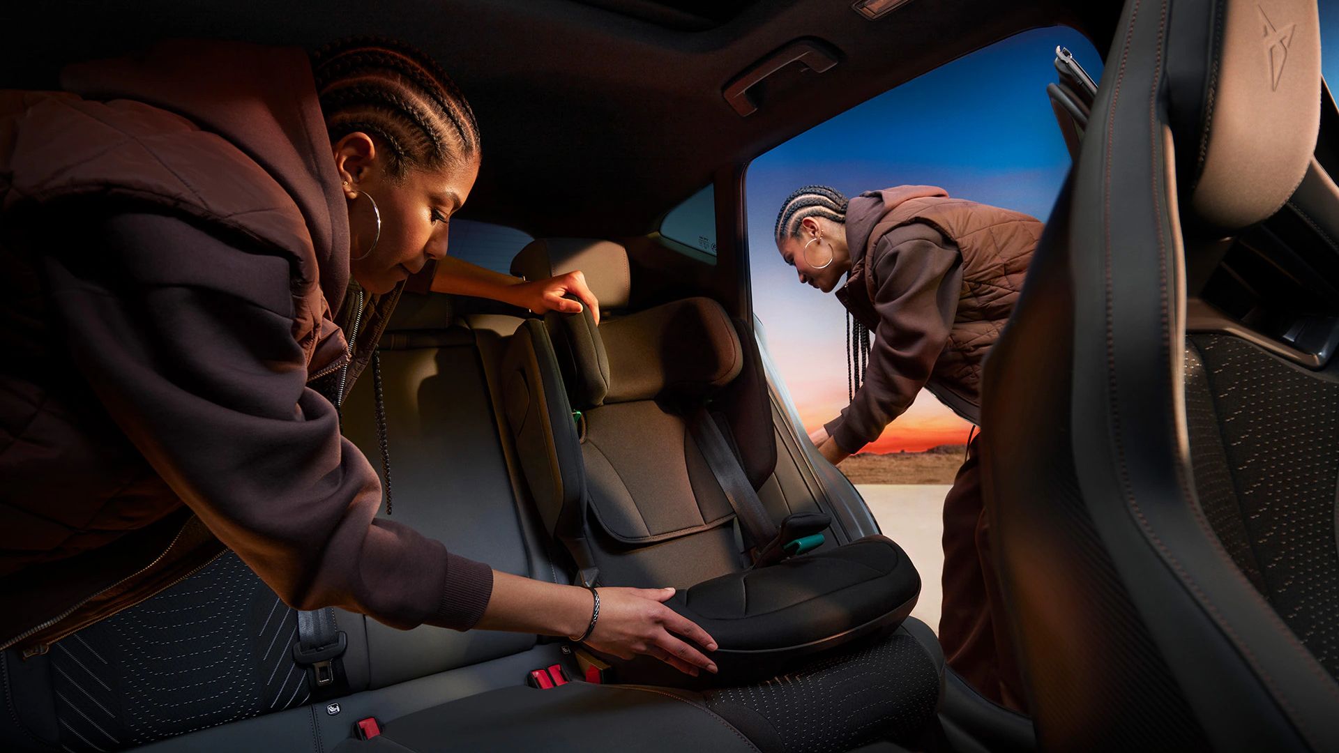 Zwei Personen befestigen einen Kindersitz in einem CUPRA Formentor. Das Innere des Autos ist sichtbar, mit aufgeklappter Rückenlehne. Im Hintergrund ist ein Sonnenuntergang zu sehen.