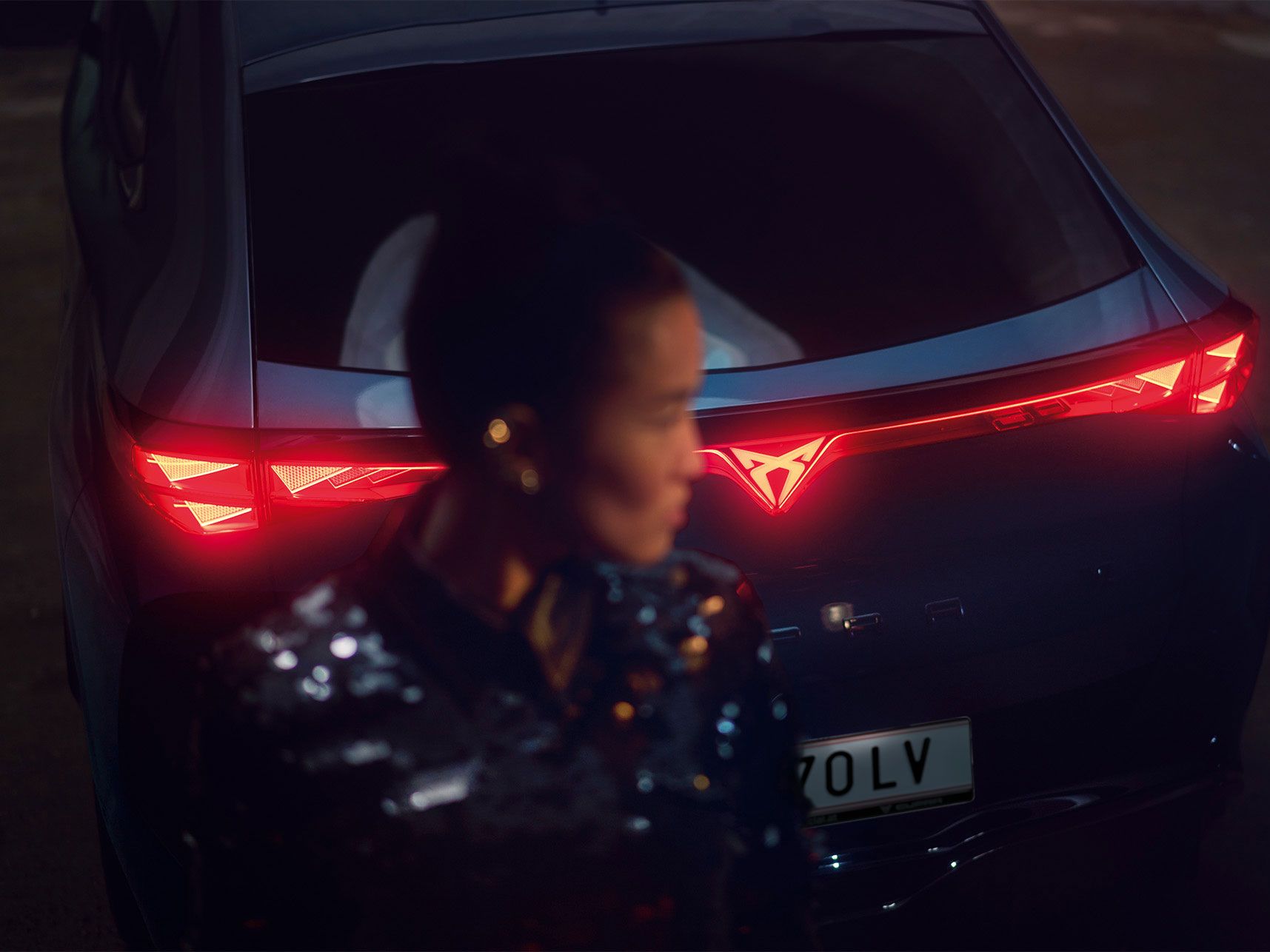 Detailaufnahme vom Heck eines CUPRA Tavascan mit durchgehender Heckbeleuchtung, eine Frau steht neben dem Fahrzeug.