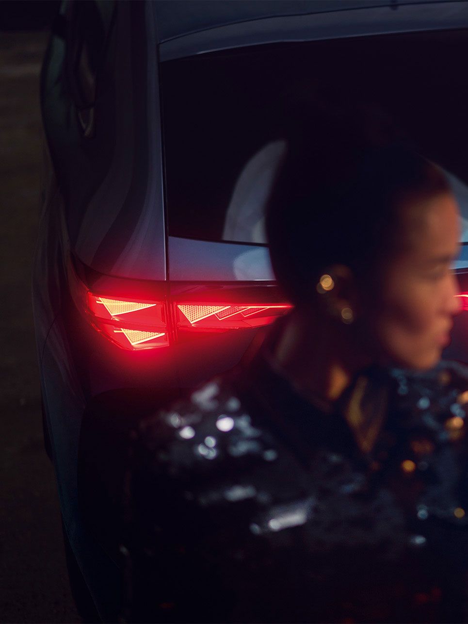 Detailaufnahme vom Heck eines CUPRA Tavascan mit durchgehender Heckbeleuchtung, eine Frau steht neben dem Fahrzeug.