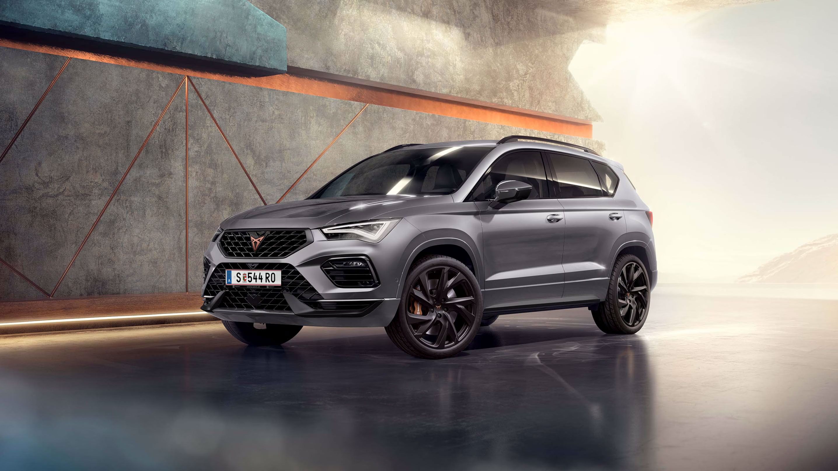 Ein 2025 Cupra Ateca in Graphitgrau, der in einem modernen, industriellen Umfeld geparkt ist. Der Hintergrund besteht aus einem geometrischen Wanddesign, das mit dem eleganten Fahrzeugdesign kontrastiert.