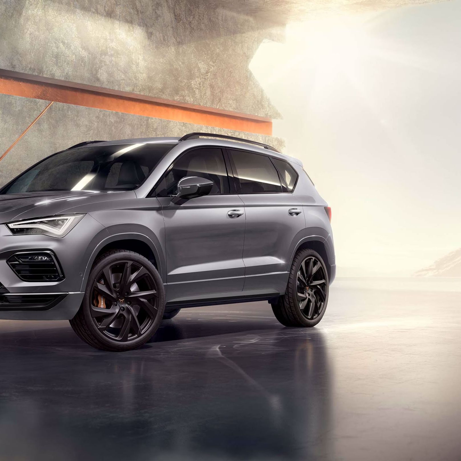 Ein 2025 Cupra Ateca in Graphitgrau, der in einem modernen, industriellen Umfeld geparkt ist. Der Hintergrund besteht aus einem geometrischen Wanddesign, das mit dem eleganten Fahrzeugdesign kontrastiert.