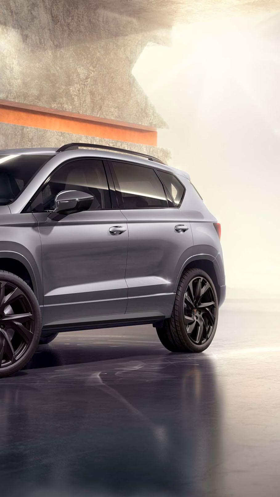 Ein 2025 Cupra Ateca in Graphitgrau, der in einem modernen, industriellen Umfeld geparkt ist. Der Hintergrund besteht aus einem geometrischen Wanddesign, das mit dem eleganten Fahrzeugdesign kontrastiert.