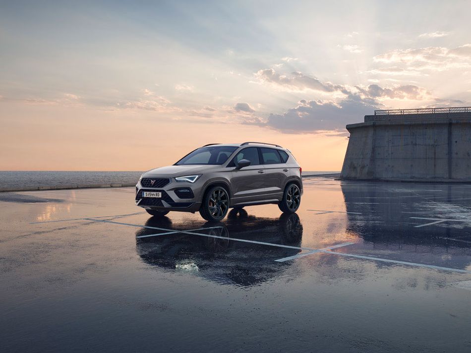 Ein grauer CUPRA Ateca parkt auf einem nassen Parkplatz an der Meeresküste.