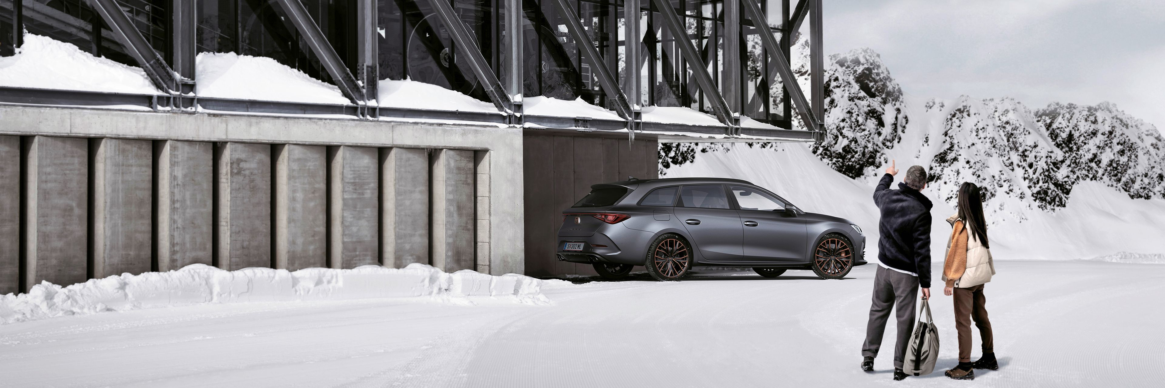 Ein grauer CUPRA Leon SP Kombi parkt vor einer Seilbahnstation in einer winterlichen Berglandschaft. Ein Mann und eine Frau blicken in Richtung des Fahrzeugs.
