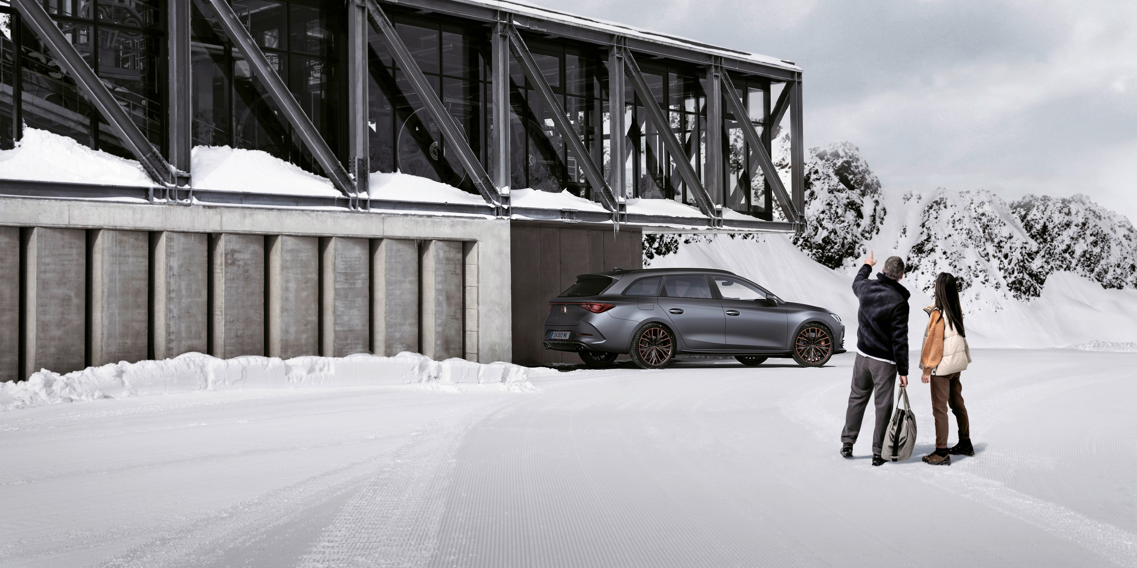 Ein grauer CUPRA Leon SP Kombi parkt vor einer Seilbahnstation in einer winterlichen Berglandschaft. Ein Mann und eine Frau blicken in Richtung des Fahrzeugs.