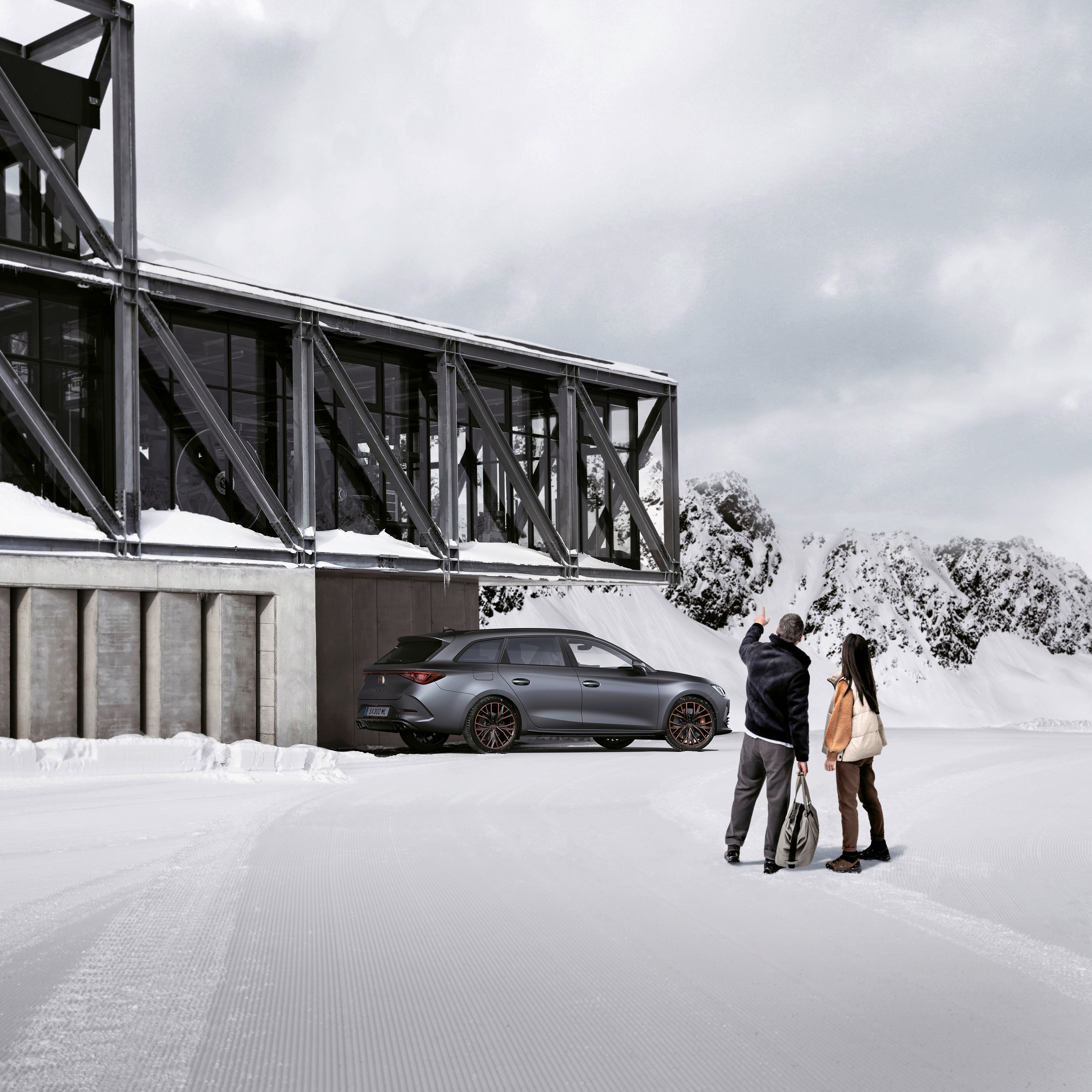 Ein grauer CUPRA Leon SP Kombi parkt vor einer Seilbahnstation in einer winterlichen Berglandschaft. Ein Mann und eine Frau blicken in Richtung des Fahrzeugs.