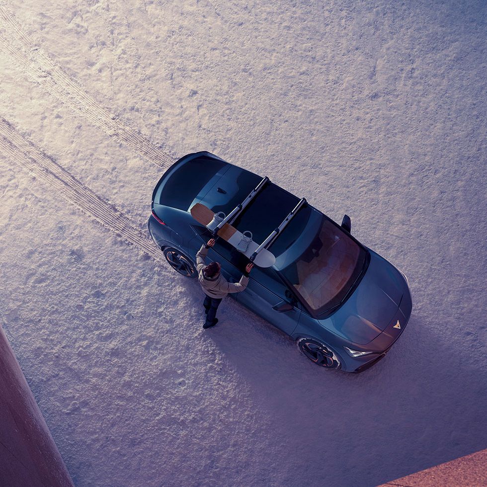 Blick von oben auf einen CUPRA Tavascan mit Snowboard Dachträger. Ein Mann montiert ein Snowboard darauf.