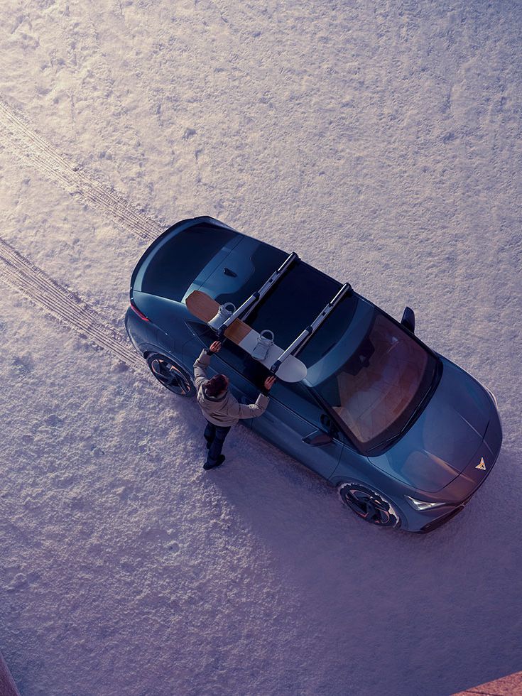 Blick von oben auf einen CUPRA Tavascan mit Snowboard Dachträger. Ein Mann montiert ein Snowboard darauf.