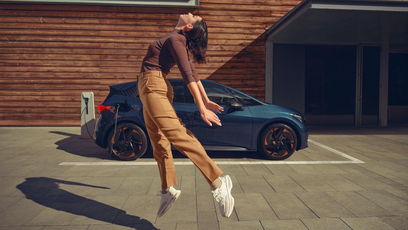 Eine Frau springt vor einem blauen CUPRA Born an einer Ladesäule in die Luft.
