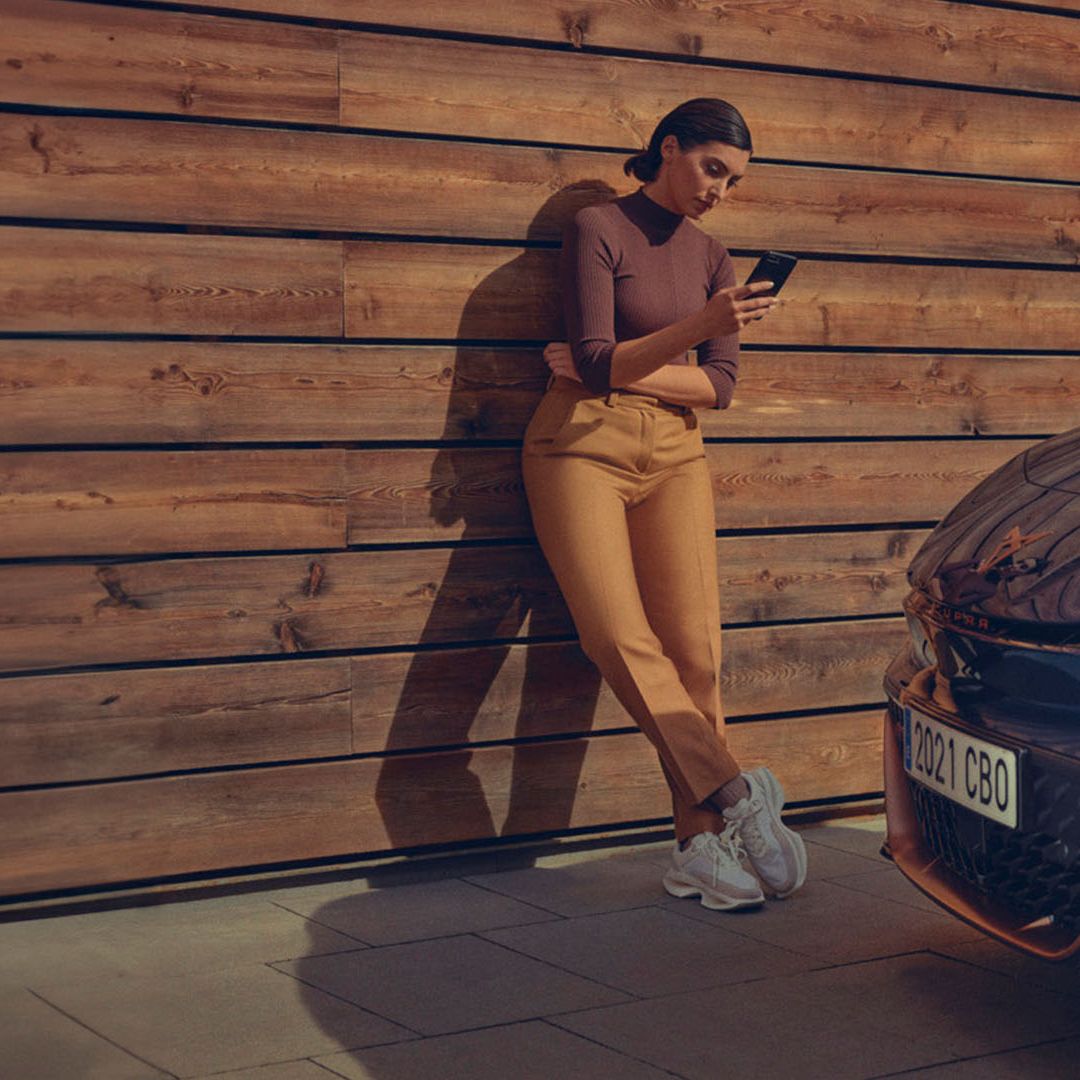 Eine Frau lehnt an einer Holzwand und blickt auf ihr Smartphone. Im Vordergrund der Kühler eines CUPRA Born.