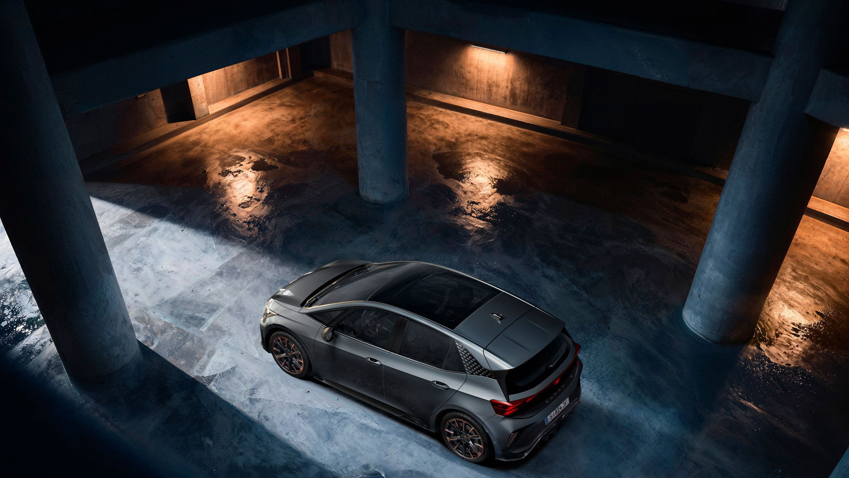Ein grauer CUPRA Born steht in einer dunklen Tiefgarage. Das Bild ist aus der Vogelperspektive aufgenommen und zeigt Lichterscheinungen auf dem nassen Betonboden. Die Szenerie ist durch indirektes Licht beleuchtet, das eine dramatische Atmosphäre schafft.