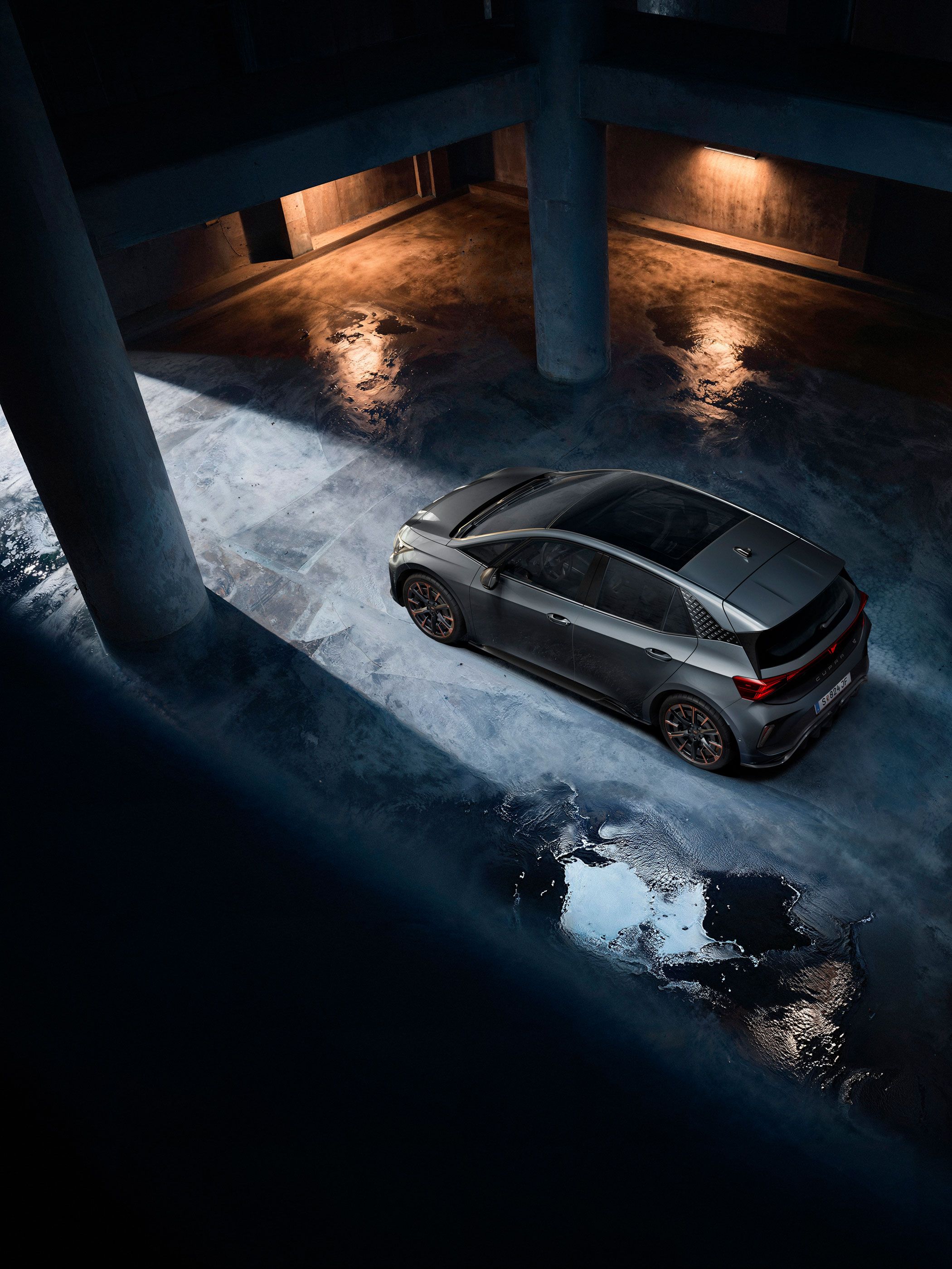 Ein grauer CUPRA Born steht in einer dunklen Tiefgarage. Das Bild ist aus der Vogelperspektive aufgenommen und zeigt Lichterscheinungen auf dem nassen Betonboden. Die Szenerie ist durch indirektes Licht beleuchtet, das eine dramatische Atmosphäre schafft.