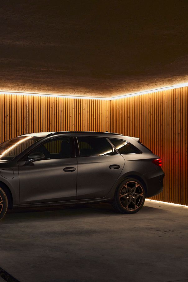 Eine Person steht in einer modernen Garage neben einem grauen CUPRA Leon SP Kombi und steckt das Ladekabel ein. Die Garage ist mit stilvollen Holzpaneelen und indirekter Beleuchtung ausgestattet. Der Boden besteht aus glattem Beton.