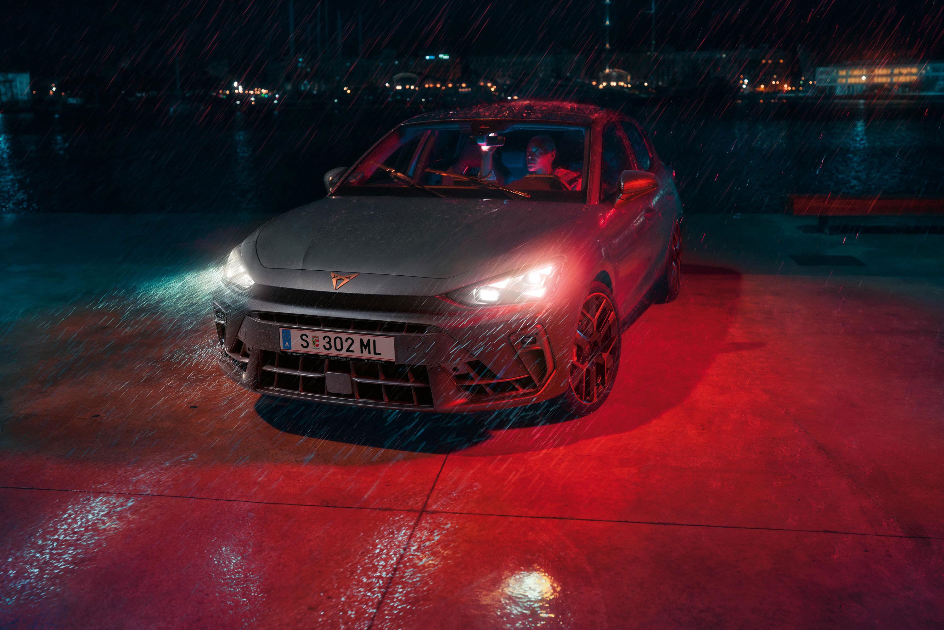 Ein Mann parkt seinen CUPRA Leon bei Nacht und Regenwetter an einem Gewässer. Im Hintergrund die Lichter einer Stadt.