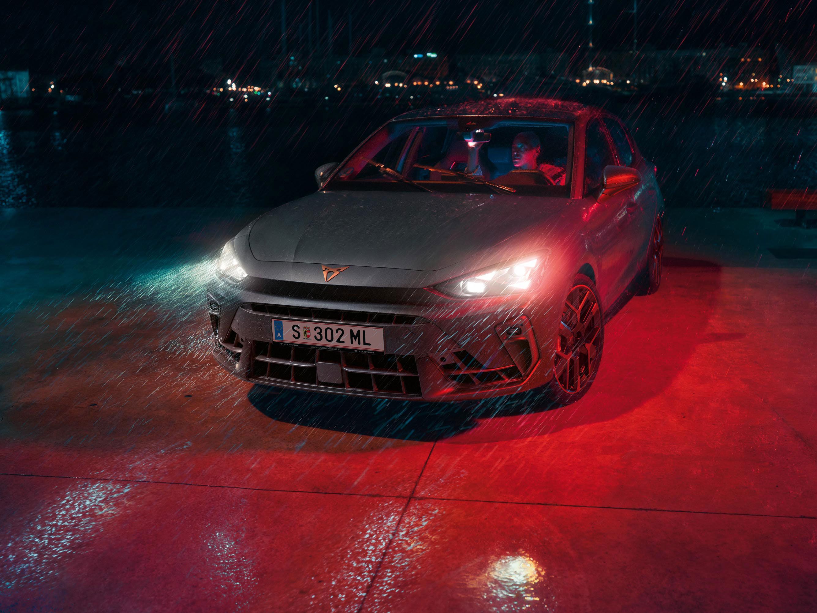 Ein Mann parkt seinen CUPRA Leon bei Nacht und Regenwetter an einem Gewässer. Im Hintergrund die Lichter einer Stadt.