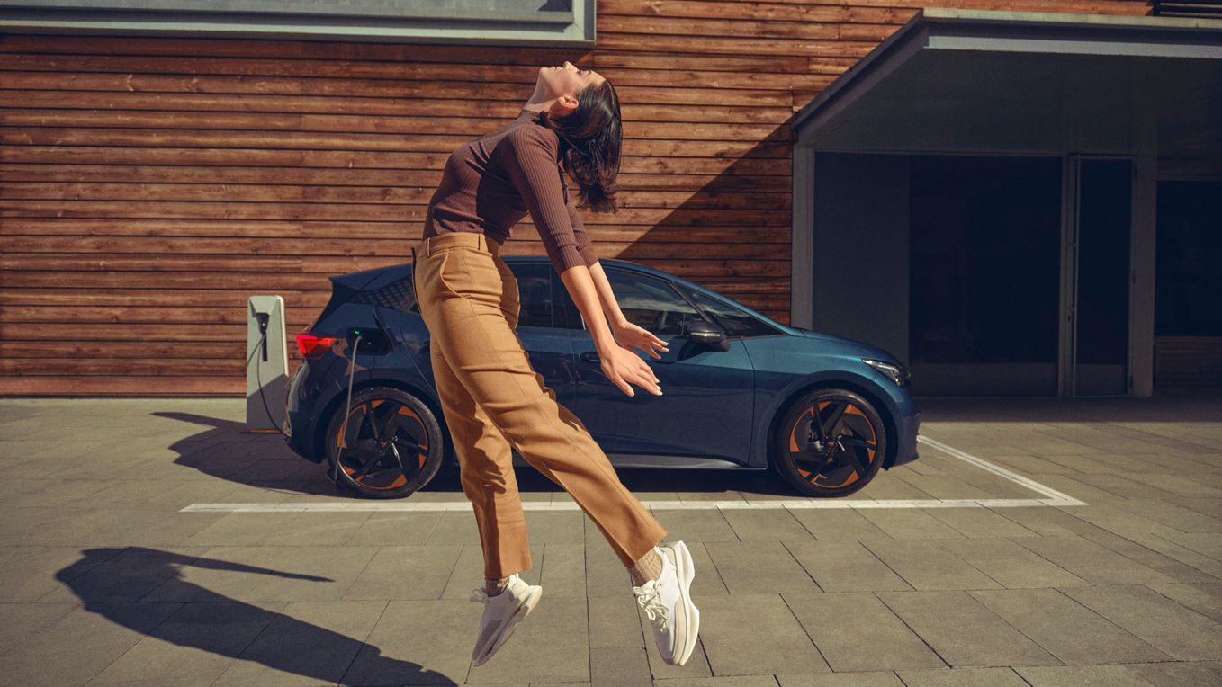 Eine Frau springt vor einem blauen CUPRA Born an einer Ladesäule in die Luft.
