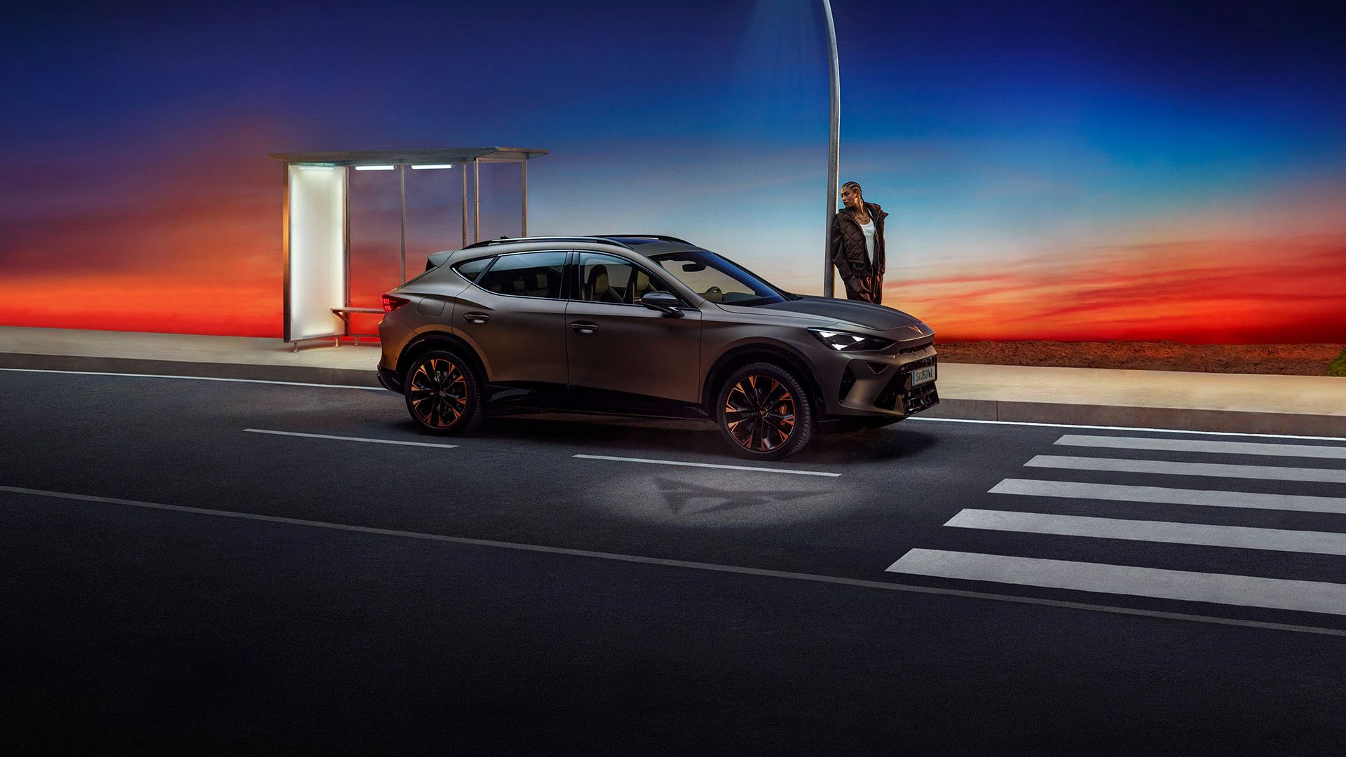Ein grauer CUPRA Formentor parkt nachts neben einer Bushaltestelle. Die Straßenlaterne beleuchtet die Szene. Am Horizont ist ein farbenfroher Sonnenuntergang zu sehen.