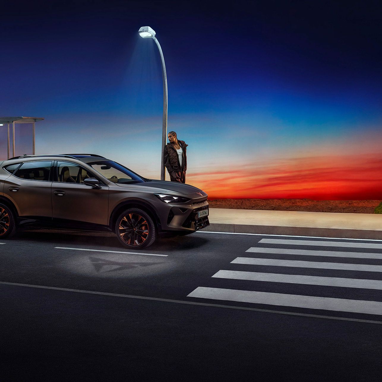 Ein grauer CUPRA Formentor parkt nachts neben einer Bushaltestelle. Die Straßenlaterne beleuchtet die Szene. Am Horizont ist ein farbenfroher Sonnenuntergang zu sehen.