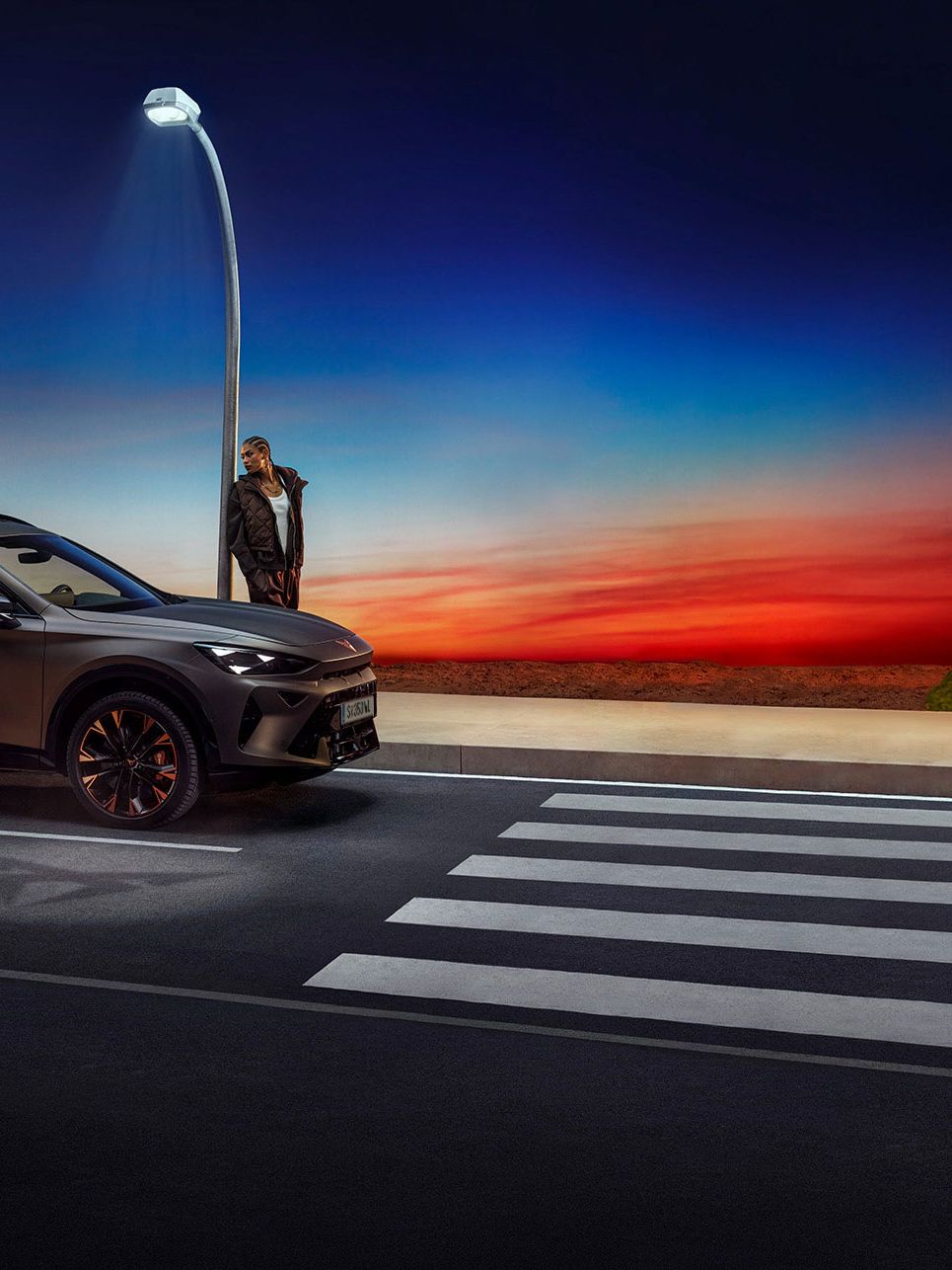 Ein grauer CUPRA Formentor parkt nachts neben einer Bushaltestelle. Die Straßenlaterne beleuchtet die Szene. Am Horizont ist ein farbenfroher Sonnenuntergang zu sehen.