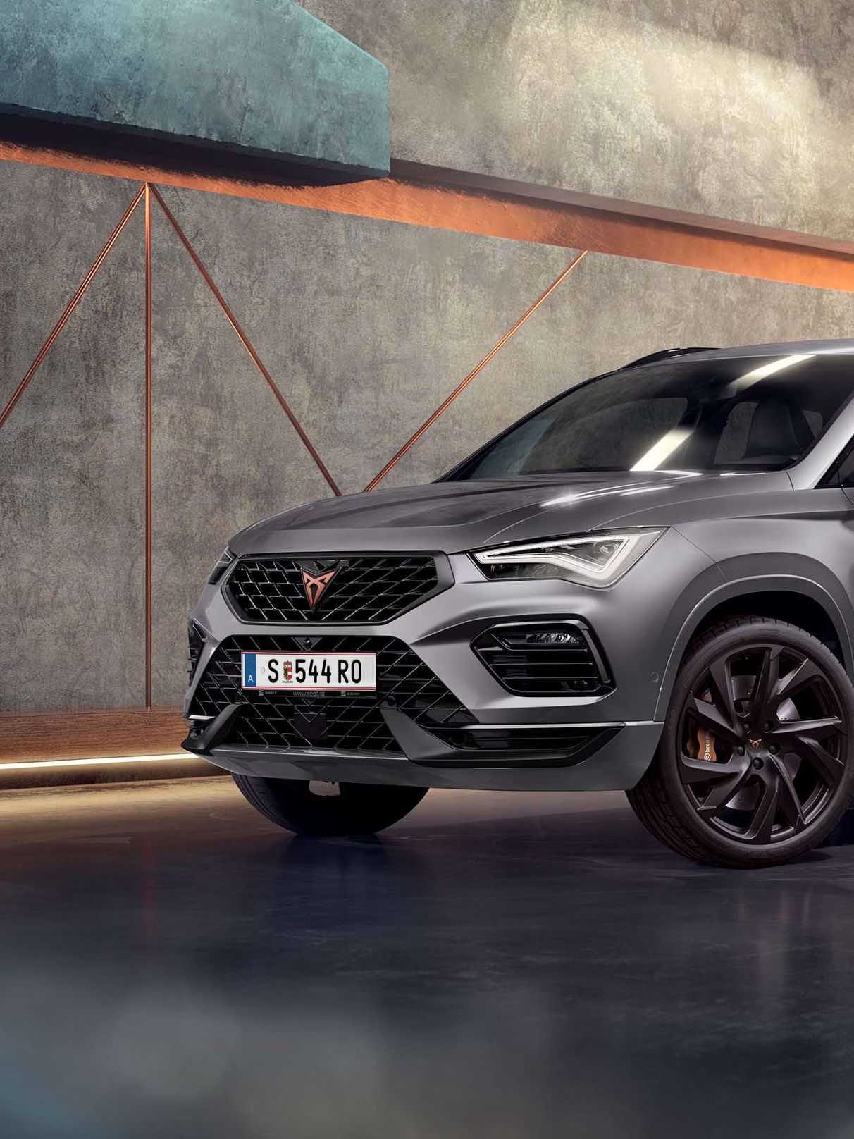 Ein 2025 Cupra Ateca in Graphitgrau, der in einem modernen, industriellen Umfeld geparkt ist. Der Hintergrund besteht aus einem geometrischen Wanddesign, das mit dem eleganten Fahrzeugdesign kontrastiert.