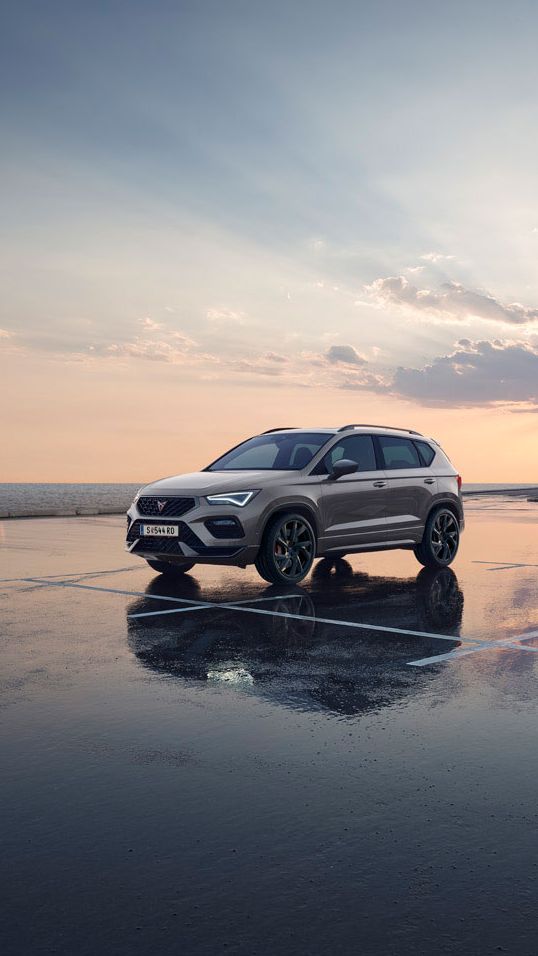 Ein grauer CUPRA Ateca parkt auf einem nassen Parkplatz an der Meeresküste.